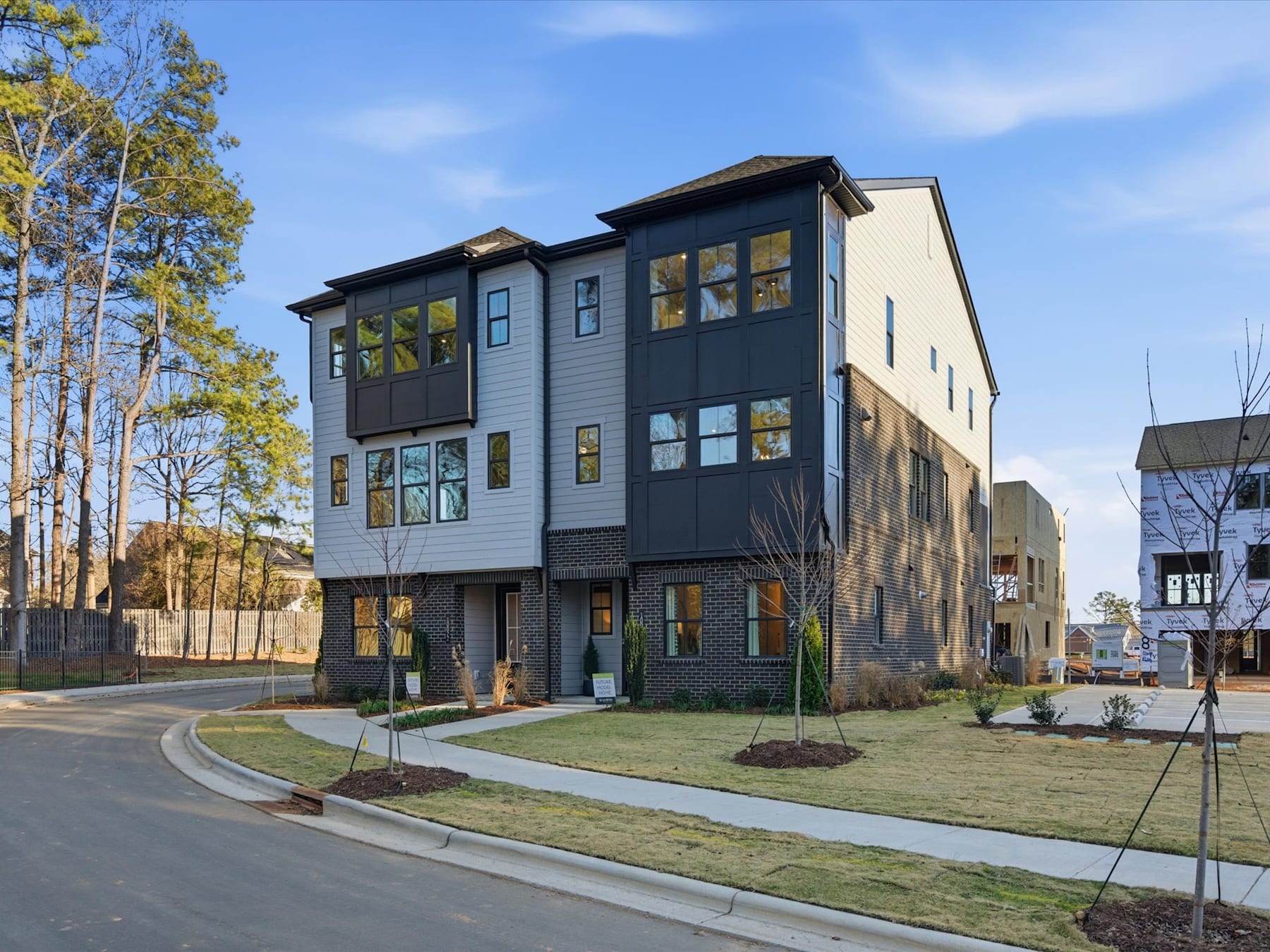 A modern multi-story residential building with a grassy lawn and landscaping in the foreground, surrounded by trees and set against a clear blue sky.