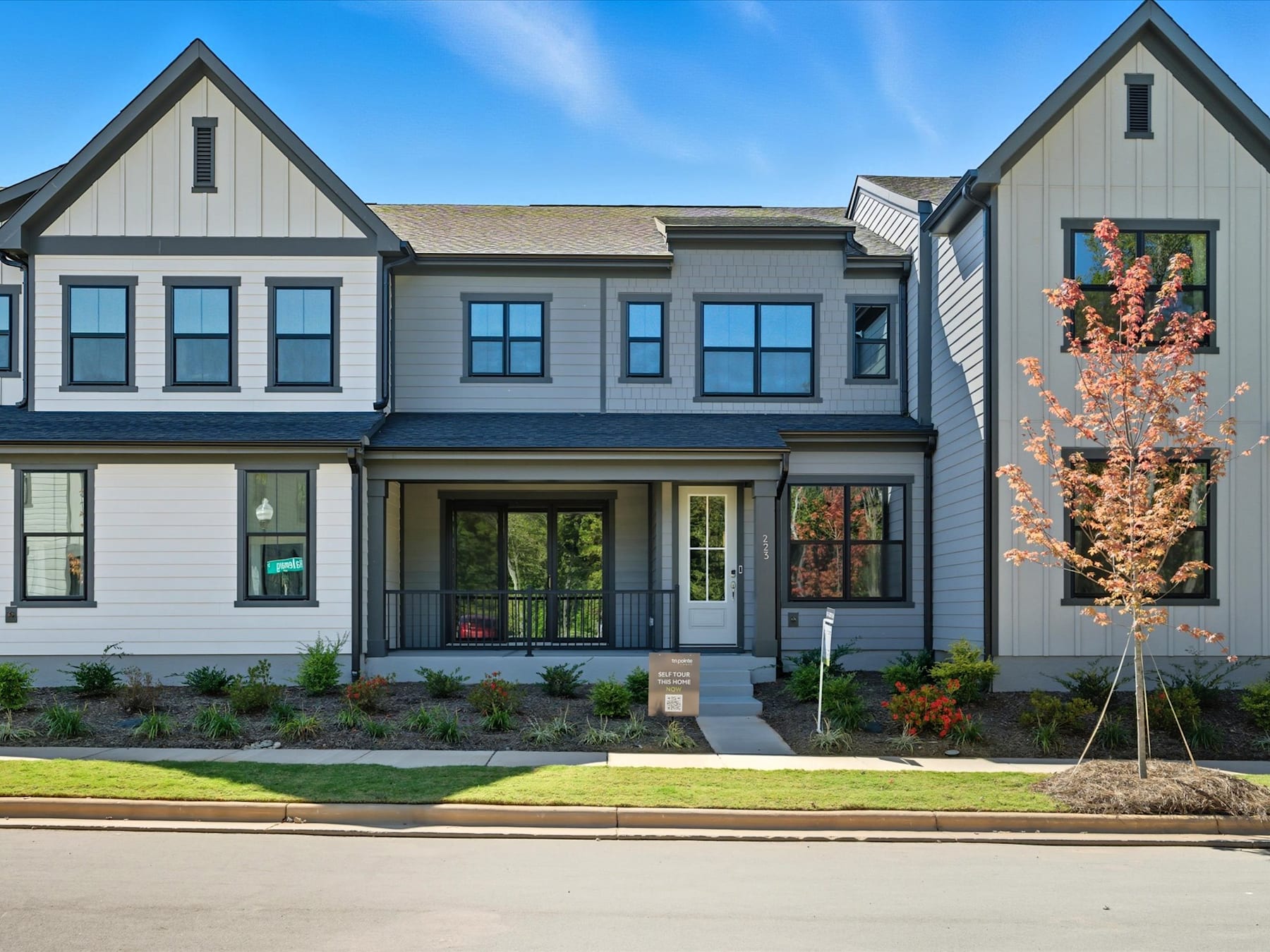 A modern, two-story townhouse with a gray exterior, black trim, and a blooming cherry tree in the foreground, set against a clear blue sky.
