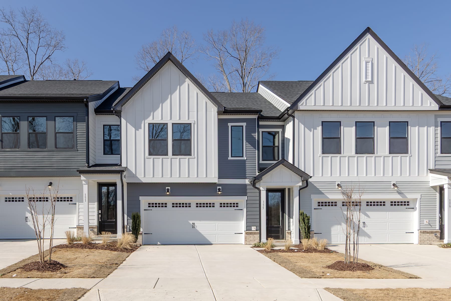 A row of modern, two-story townhouses with white siding, black roofs, and attached garages, set against a backdrop of bare trees and a clear blue sky.