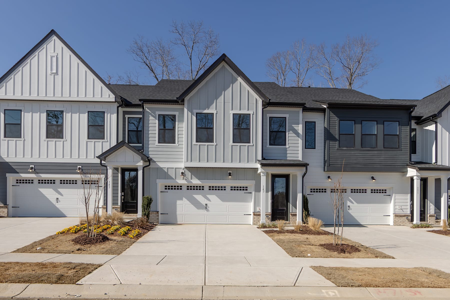 A row of modern, two-story townhouses with white siding, black roofs, and attached garages, set against a backdrop of bare trees and a clear blue sky.