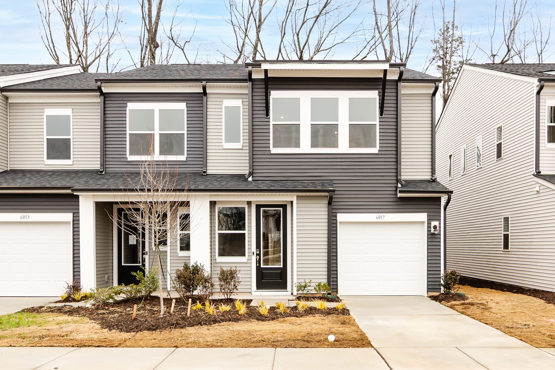 A two-story townhouse with a garage, surrounded by bare trees and a grassy yard in the foreground.