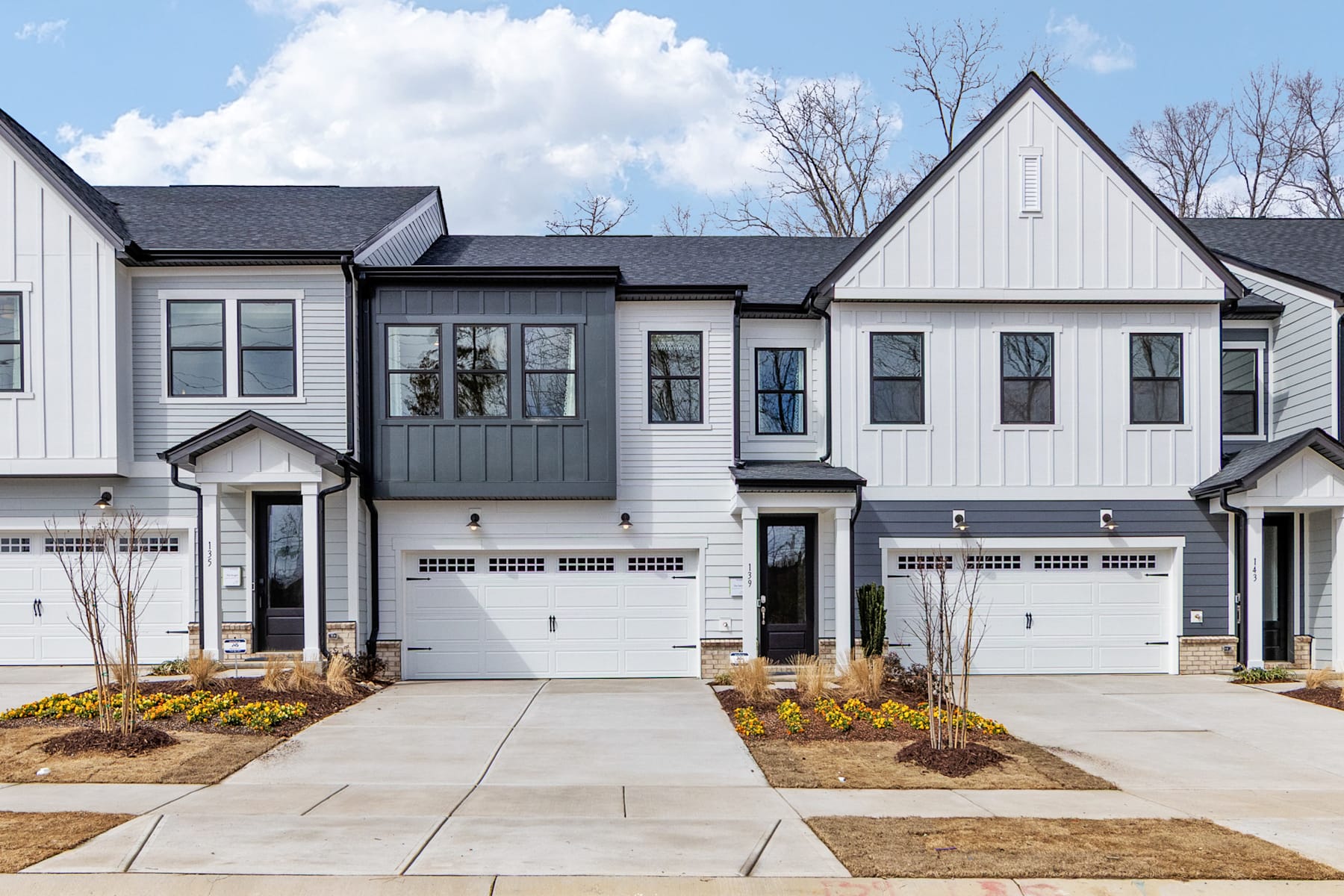 A row of modern, two-story townhouses with white siding, gray accents, and attached garages, set against a backdrop of bare trees and a cloudy sky.