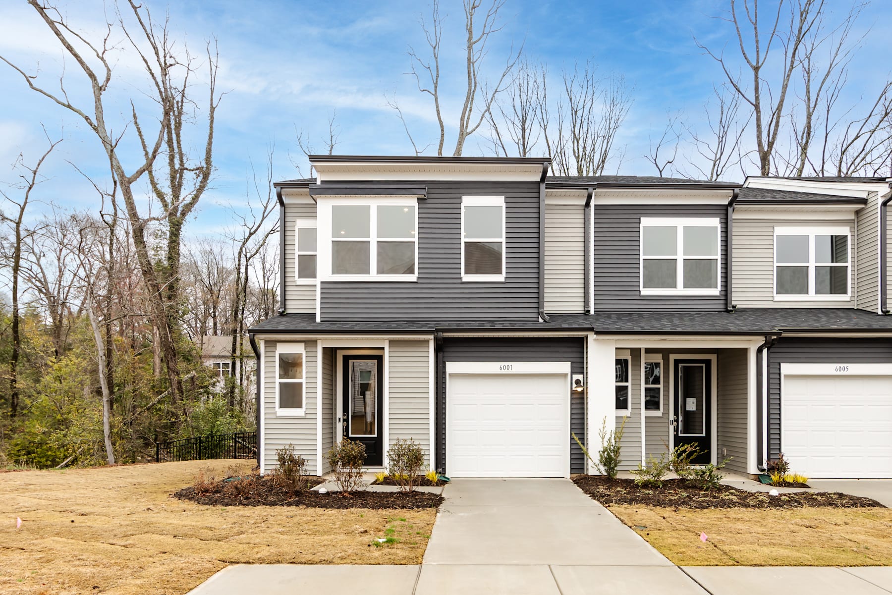 A modern two-story townhouse with gray siding and white trim stands in a wooded area, surrounded by bare trees and a grassy lawn in the foreground.