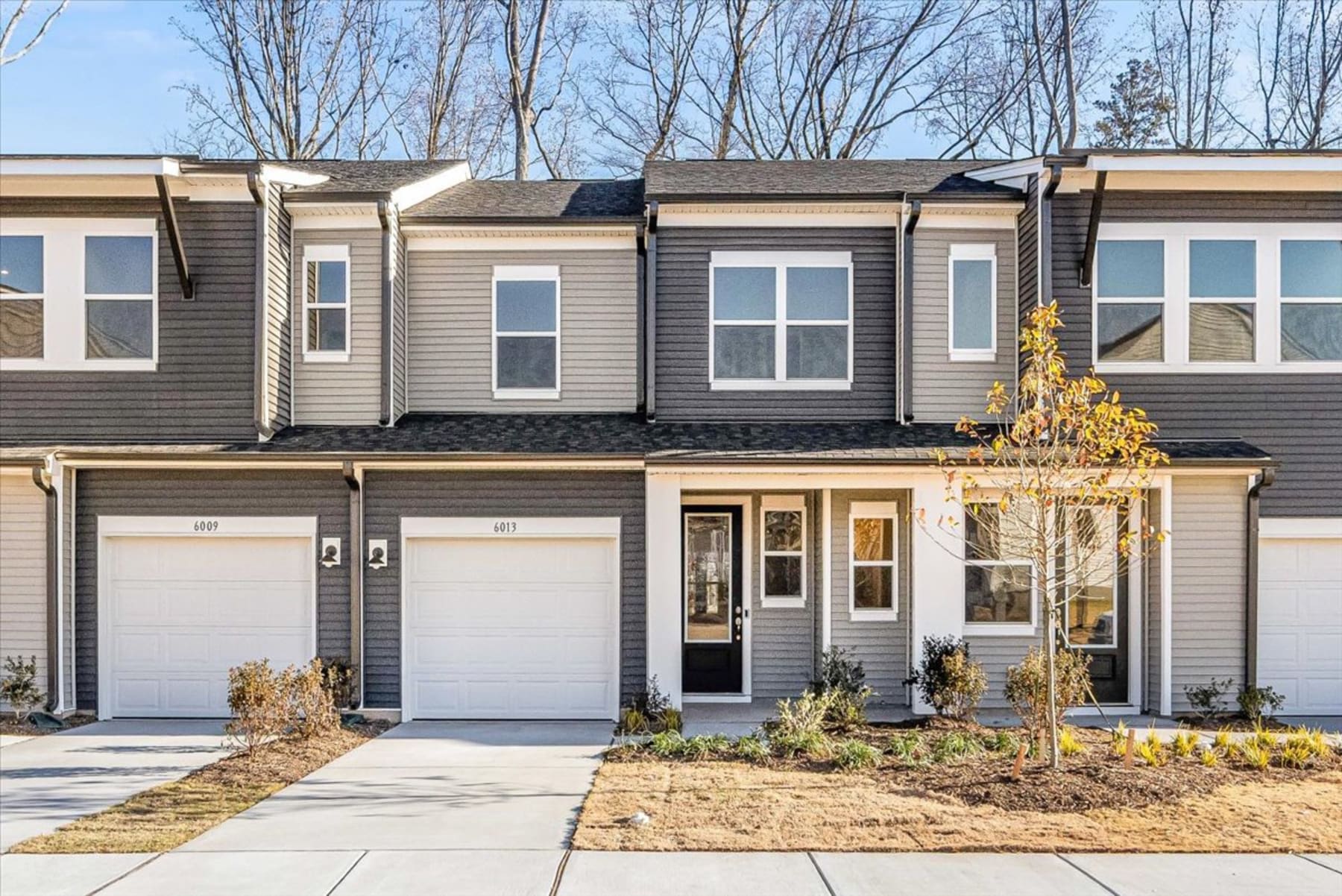 A modern townhouse with a garage, surrounded by bare trees and a landscaped yard in the foreground.