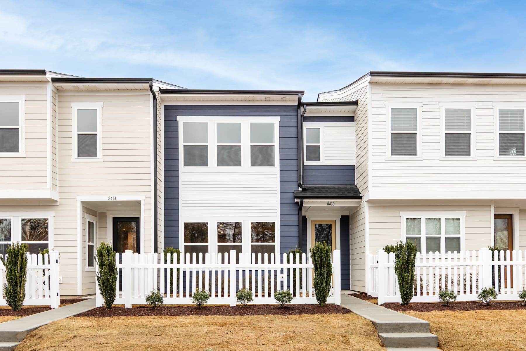 A row of modern, two-story townhouses with white siding, black trim, and white picket fences in the foreground, set against a clear blue sky in the background.