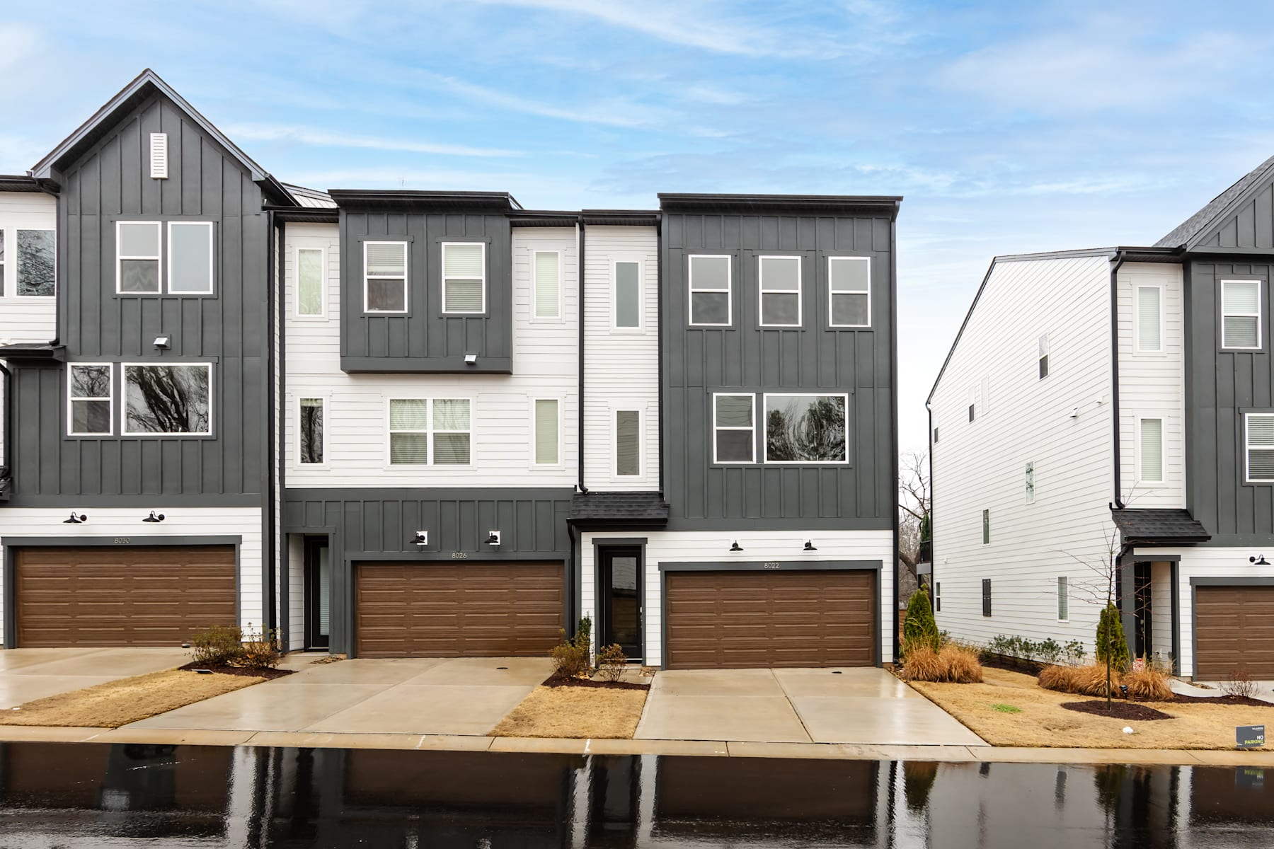 A row of modern, multi-story townhouses with gray siding, large windows, and attached garages, set against a blue sky with wispy clouds.