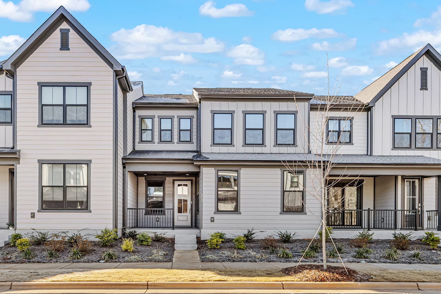 A row of modern, gray-colored townhouses with gabled roofs and large windows, set against a backdrop of a blue sky with fluffy white clouds.