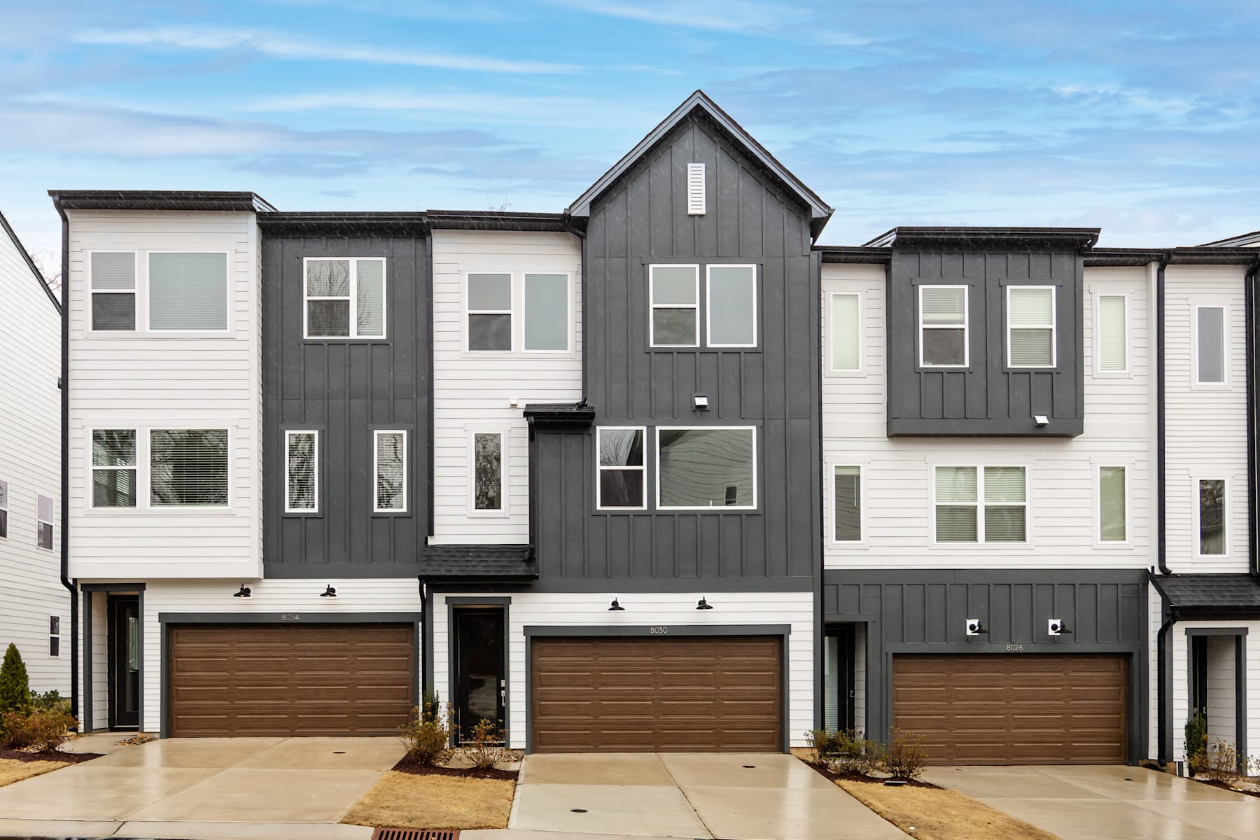 The image depicts a modern, multi-story townhouse complex with a mix of light and dark exterior colors, featuring garages and large windows against a backdrop of a blue sky with wispy clouds.