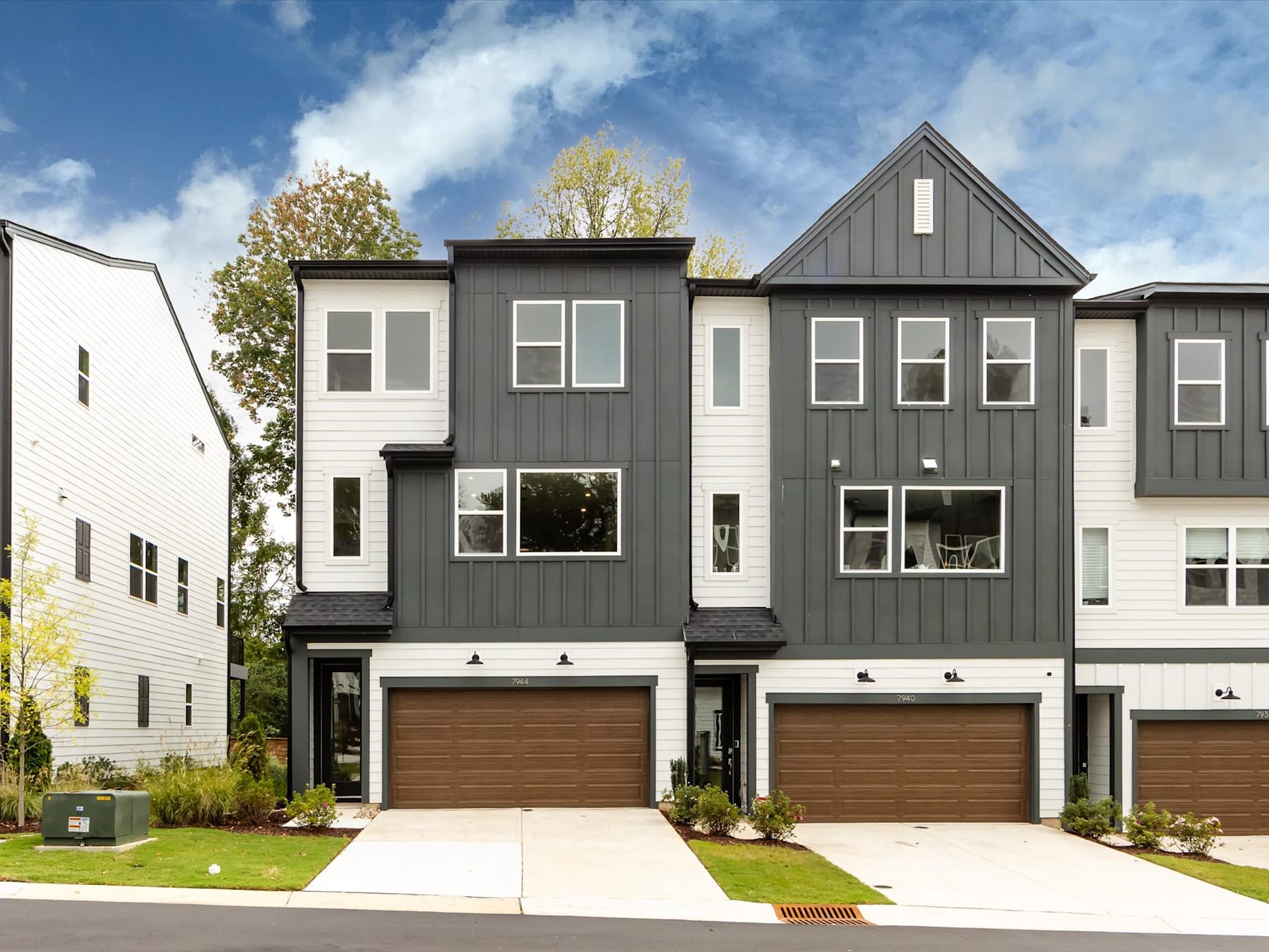 The image depicts a modern, multi-story townhouse complex with a mix of gray and white siding, large windows, and attached garages. The townhouses are set against a backdrop of blue sky with scattered clouds, and the foreground features a grassy lawn and paved walkway.