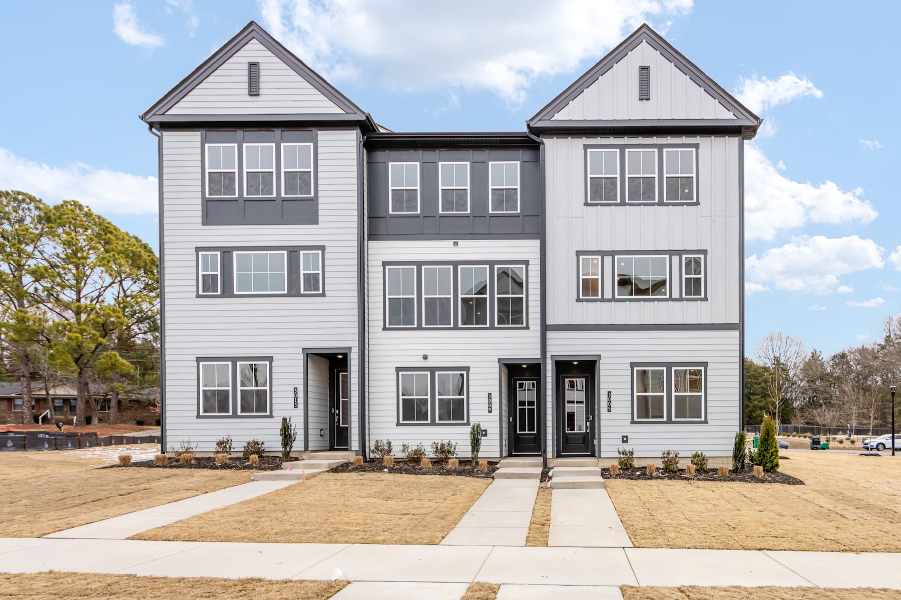 A modern, multi-unit residential building with a gray exterior and gabled roofs, surrounded by a paved walkway and landscaping in the foreground.