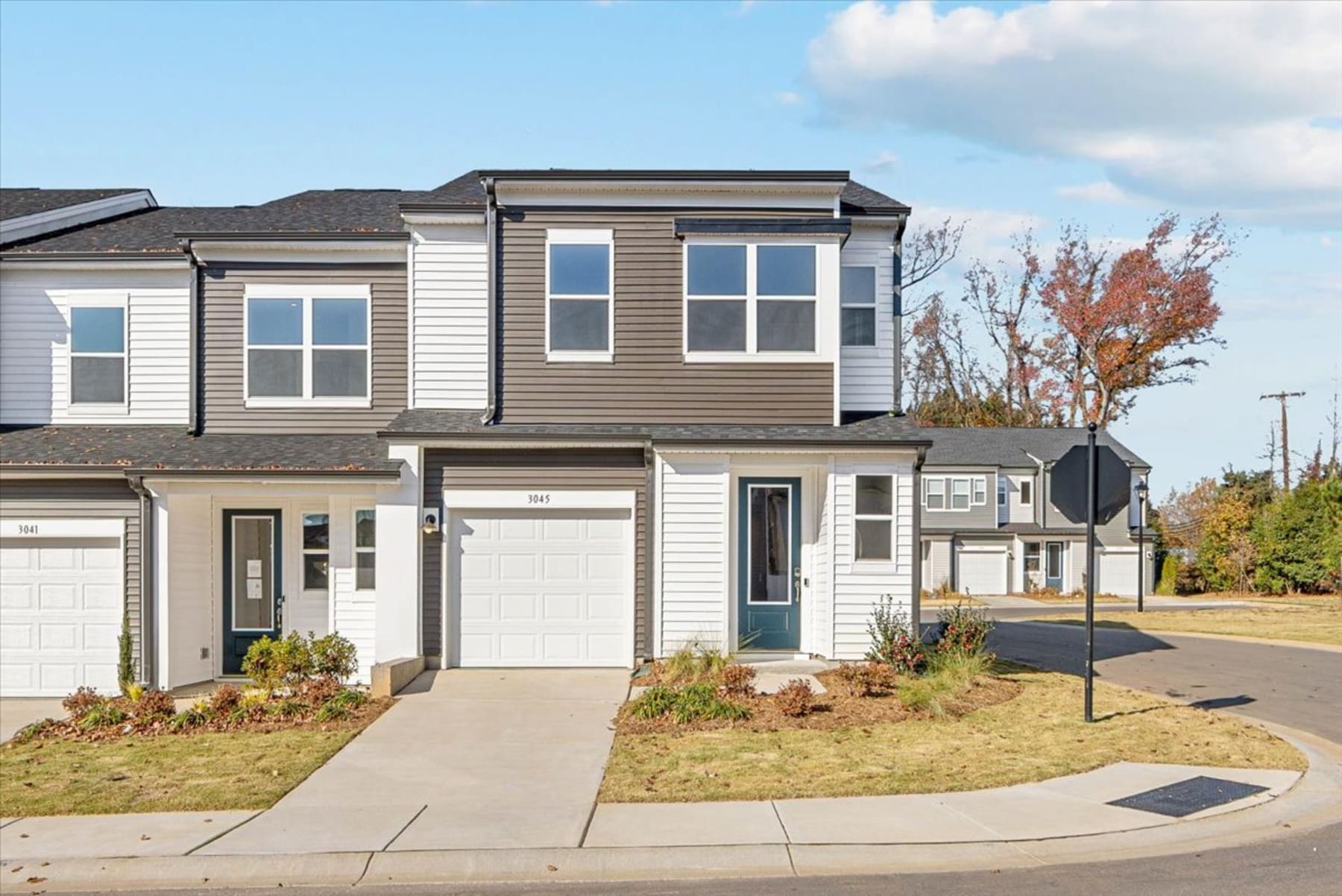 The image depicts a modern townhouse with a two-car garage, surrounded by a well-maintained lawn and landscaping, set against a backdrop of autumn foliage.