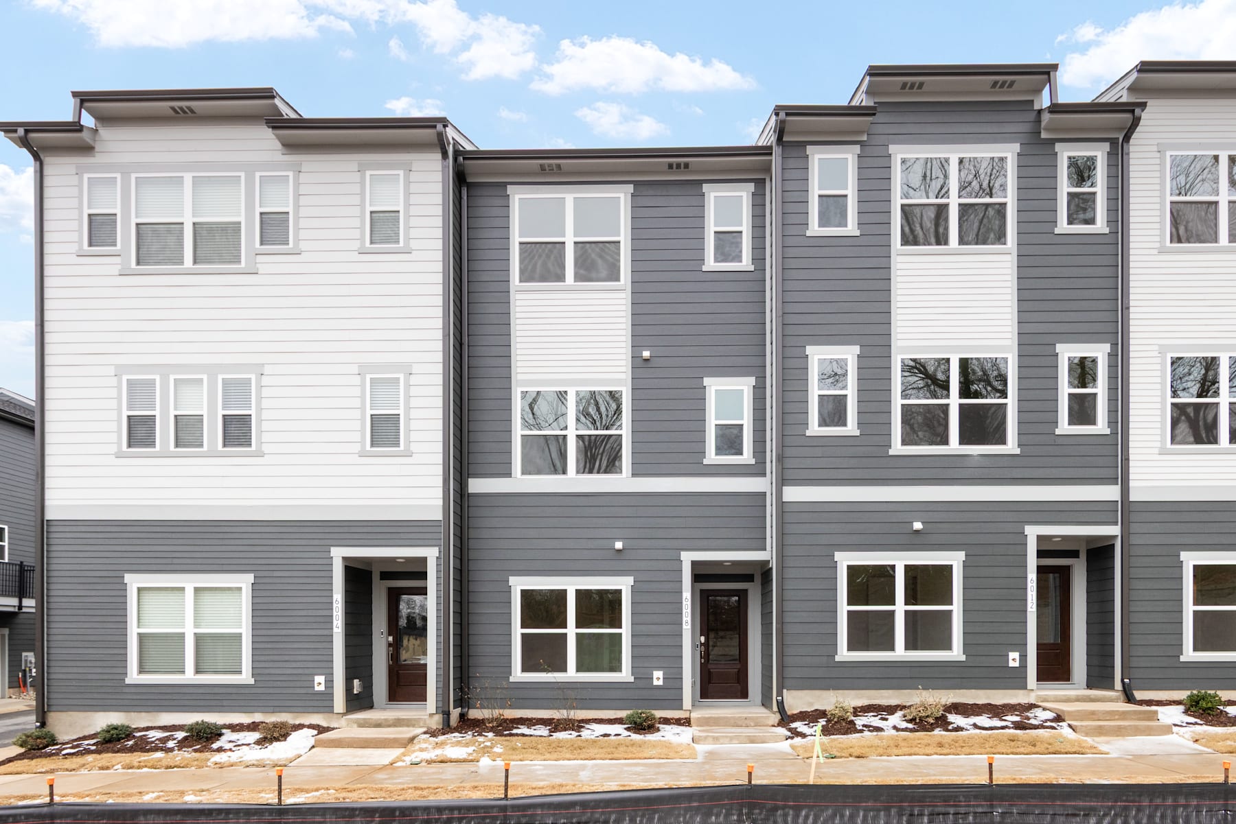The image depicts a row of modern, multi-story townhouses with a mix of gray and white siding, featuring numerous windows and decorative architectural elements. The foreground includes landscaping with rocks and plants, while the background shows a partly cloudy sky.