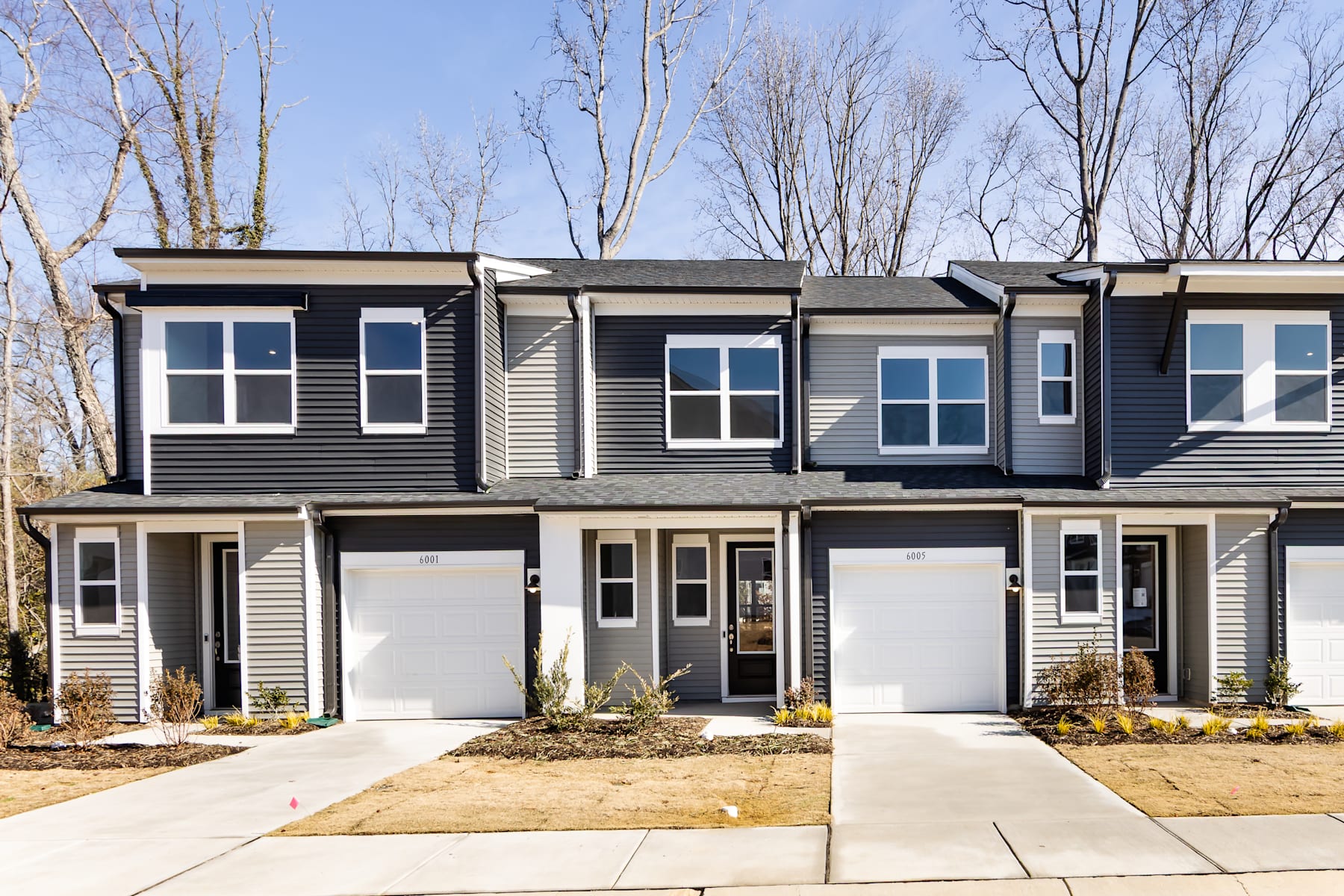The image depicts a row of modern townhouses with gray siding and white trim, set against a backdrop of bare trees and a clear blue sky.