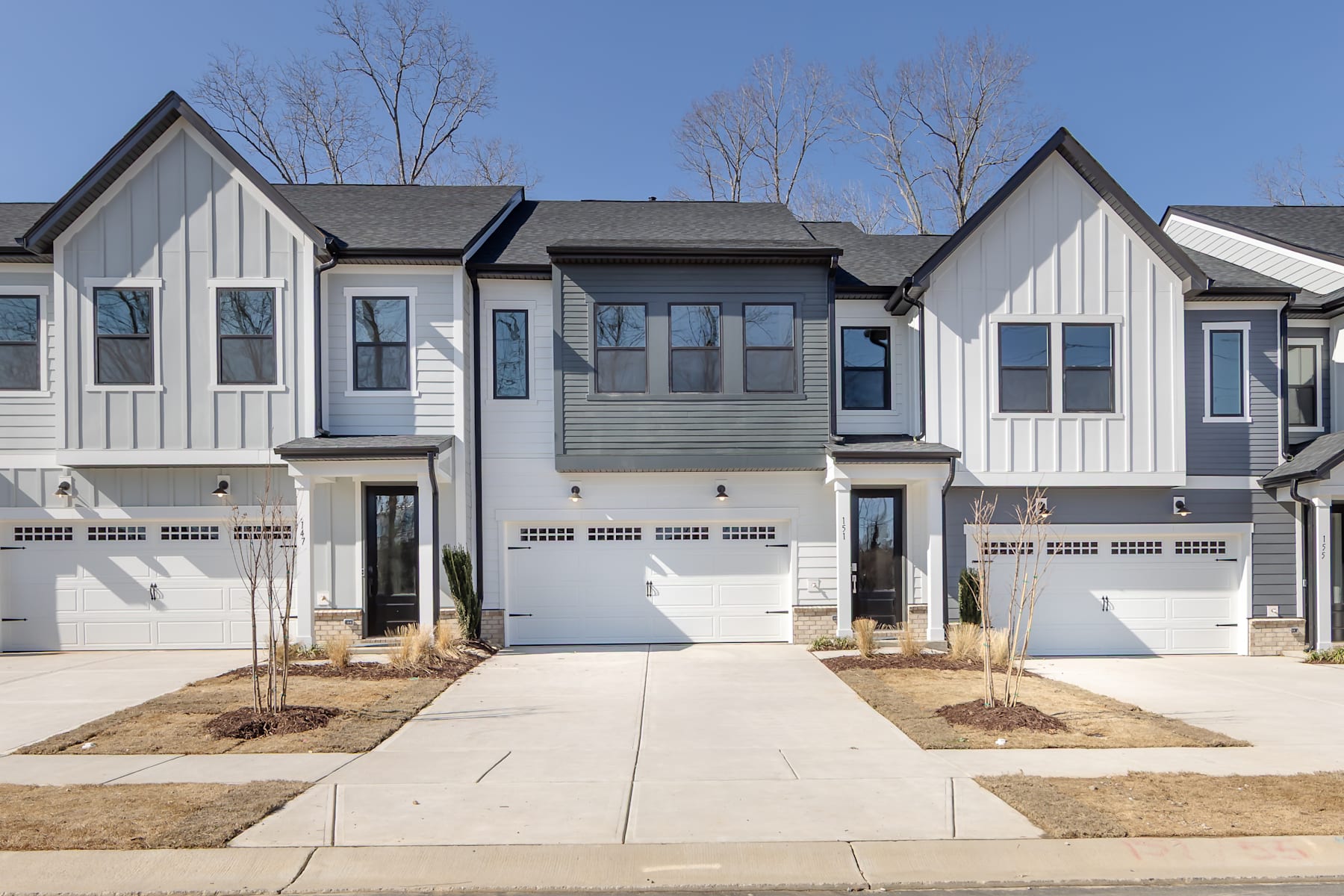 The image depicts a row of modern townhouses with white siding, black roofs, and attached garages. The townhouses are set against a backdrop of bare trees and a clear blue sky.