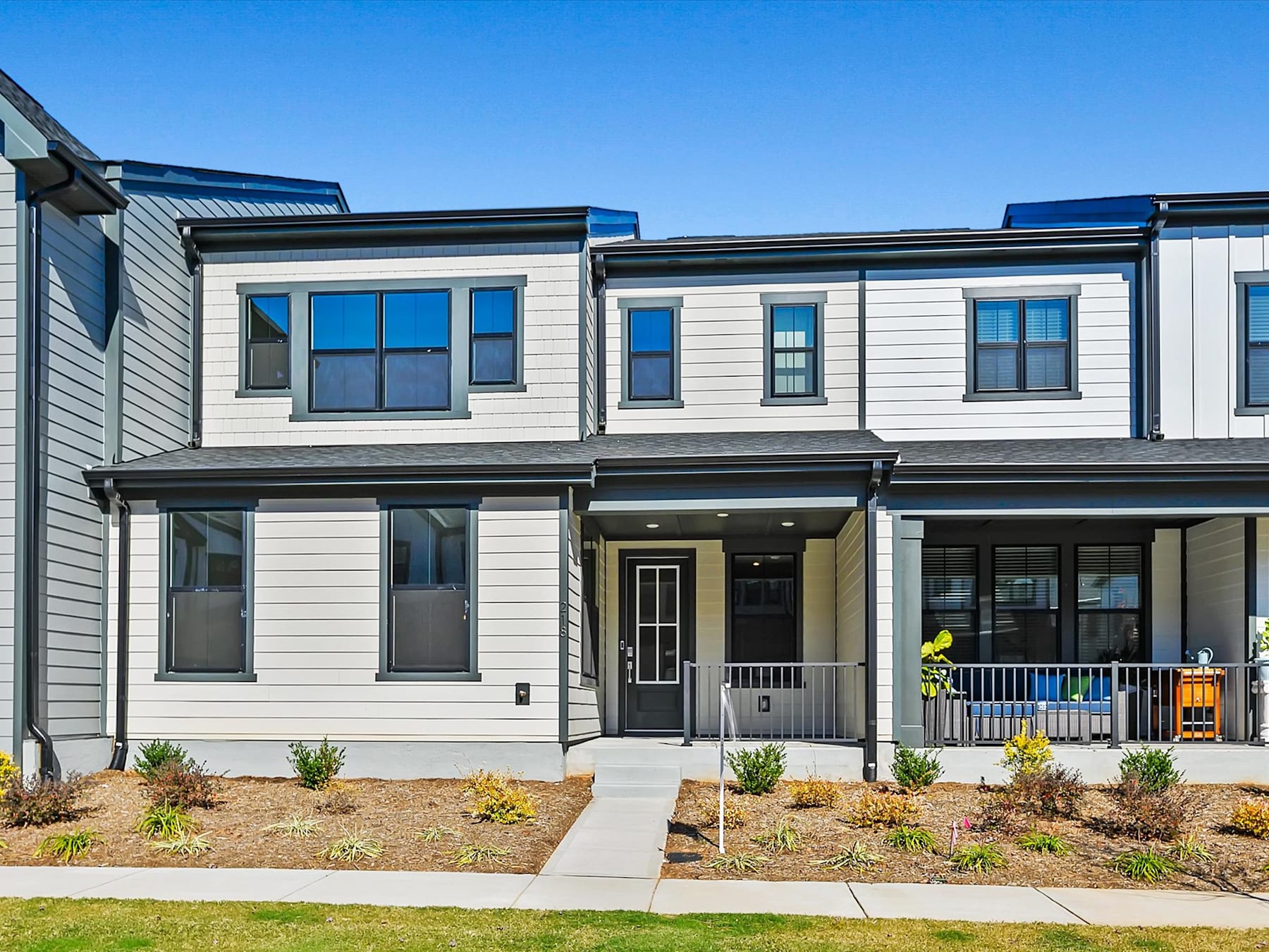 A modern, multi-story townhouse with a porch and landscaped yard, set against a clear blue sky.