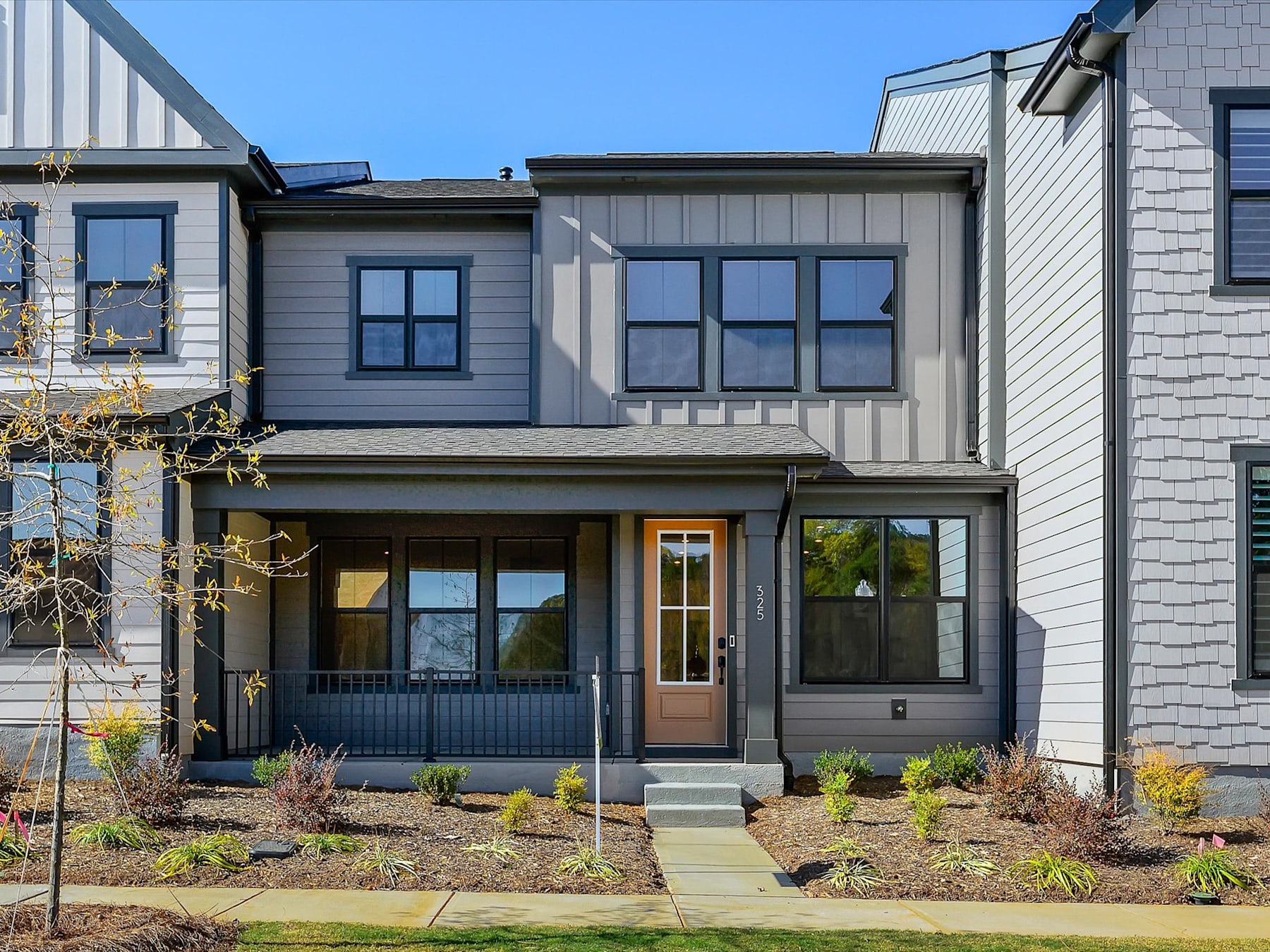 A modern, two-story townhouse with a wooden front door, surrounded by landscaped greenery and set against a clear blue sky.