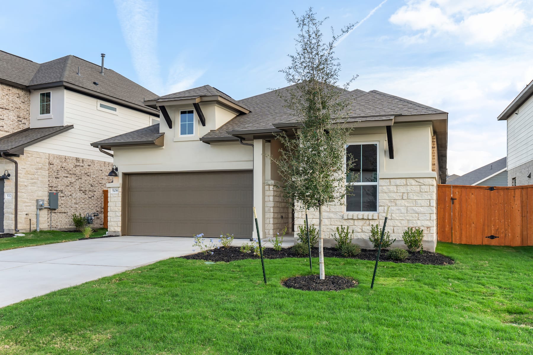 A newly constructed two-story house with a well-manicured lawn and a young tree in the foreground, set against a clear blue sky with scattered clouds.