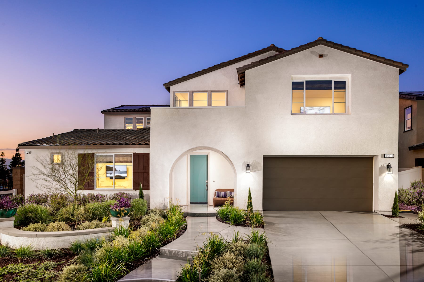 A modern, two-story white stucco house with a tiled roof, surrounded by a well-landscaped yard featuring a paved driveway, steps, and various plants and shrubs, set against a clear blue sky at sunset.