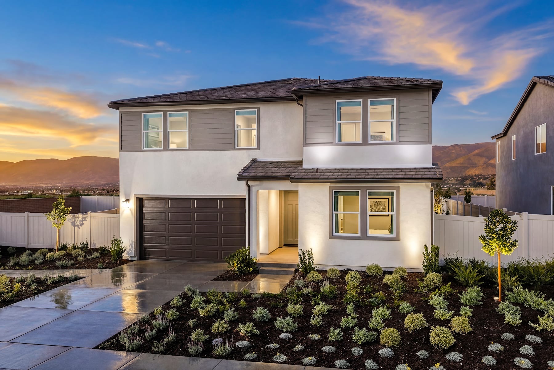 A modern two-story house with a garage, surrounded by a well-landscaped yard featuring a variety of plants and a paved driveway, set against a vibrant sunset sky.