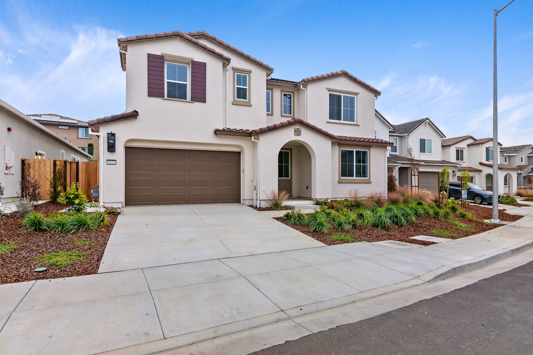 A two-story Mediterranean-style house with a garage, surrounded by a well-landscaped yard and a paved driveway, set against a clear blue sky.