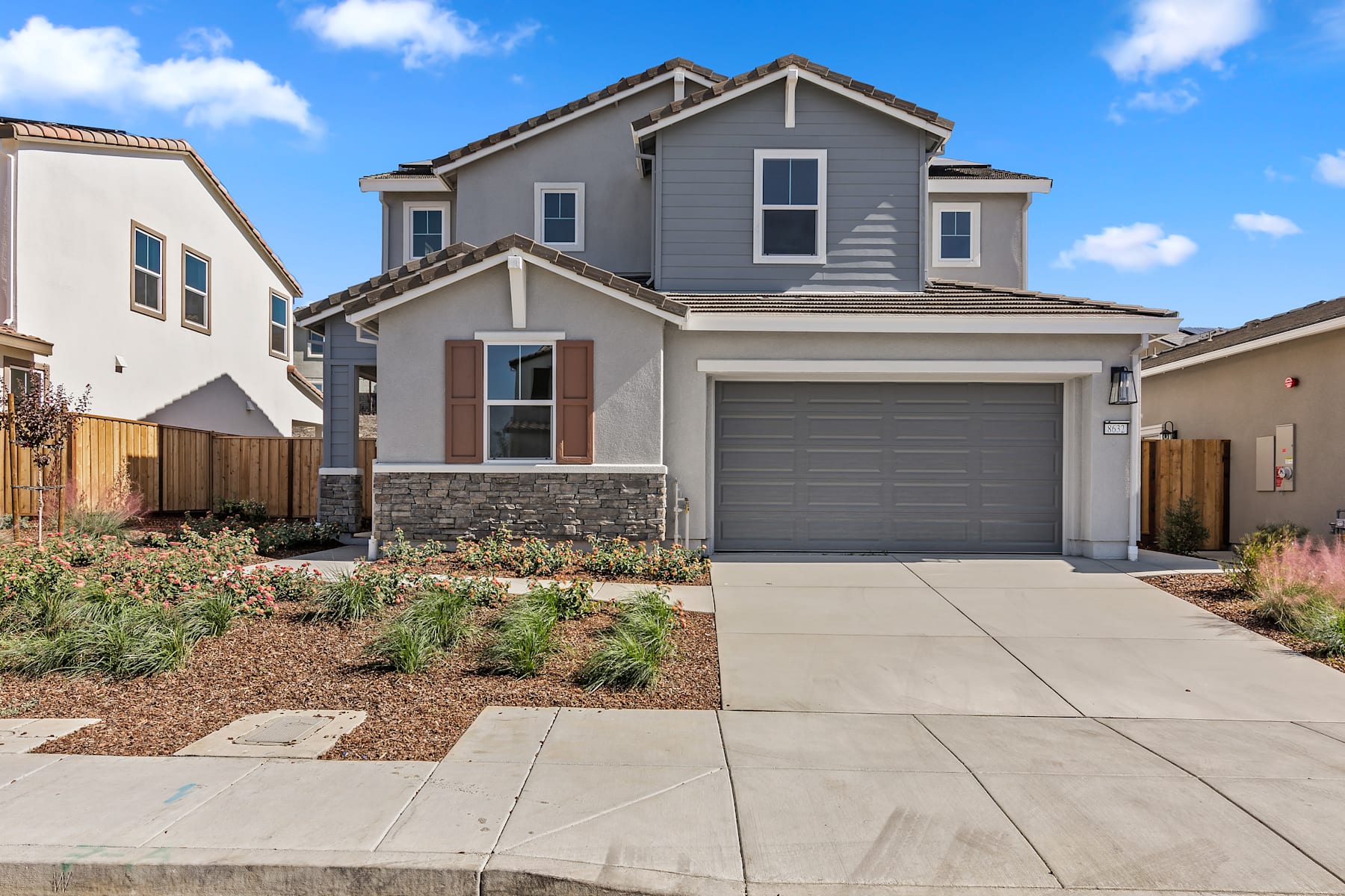 A two-story gray house with a garage, surrounded by a paved driveway and landscaped yard, set against a blue sky with fluffy white clouds.