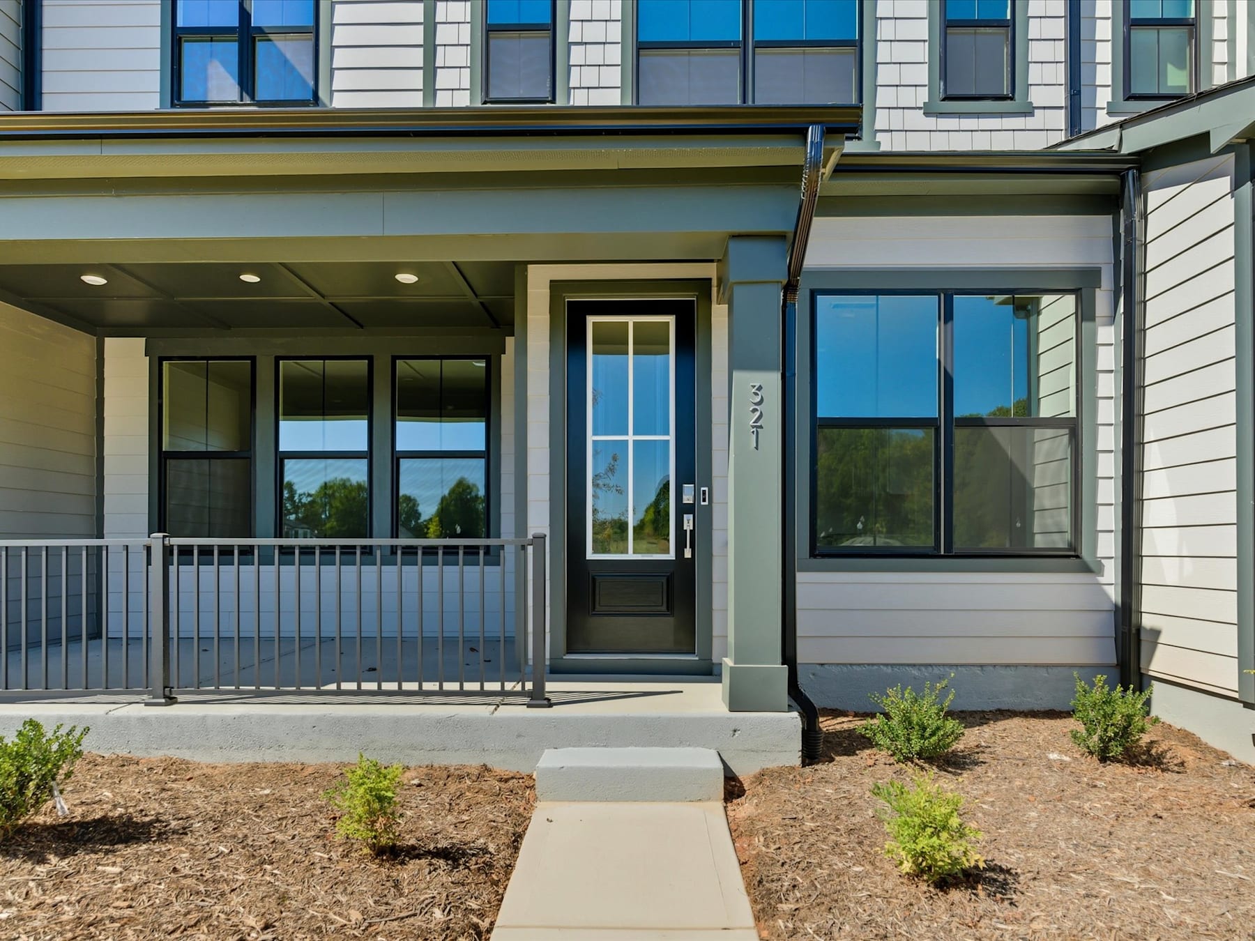 A modern, two-story residential building with a covered entryway, surrounded by newly planted landscaping and a paved walkway leading to the front door.