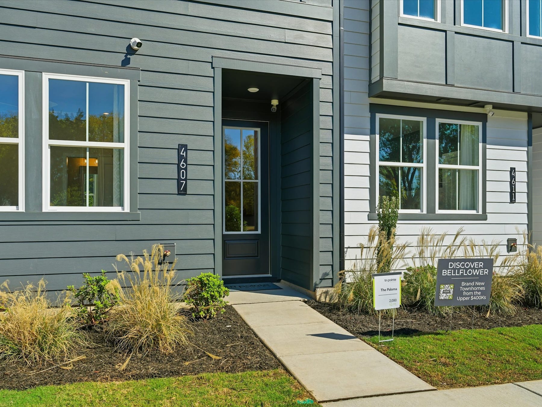 The image shows the exterior of a modern, gray-colored building with large windows and a prominent entrance. In the foreground, there is a paved walkway leading to the entrance, surrounded by landscaping with plants and a sign indicating "Discover Bellevue".