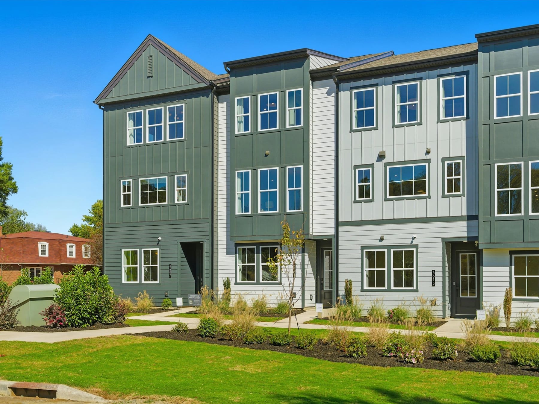 The image depicts a row of modern, multi-story townhouses with green siding and large windows, set against a clear blue sky and surrounded by a well-manicured lawn in the foreground.