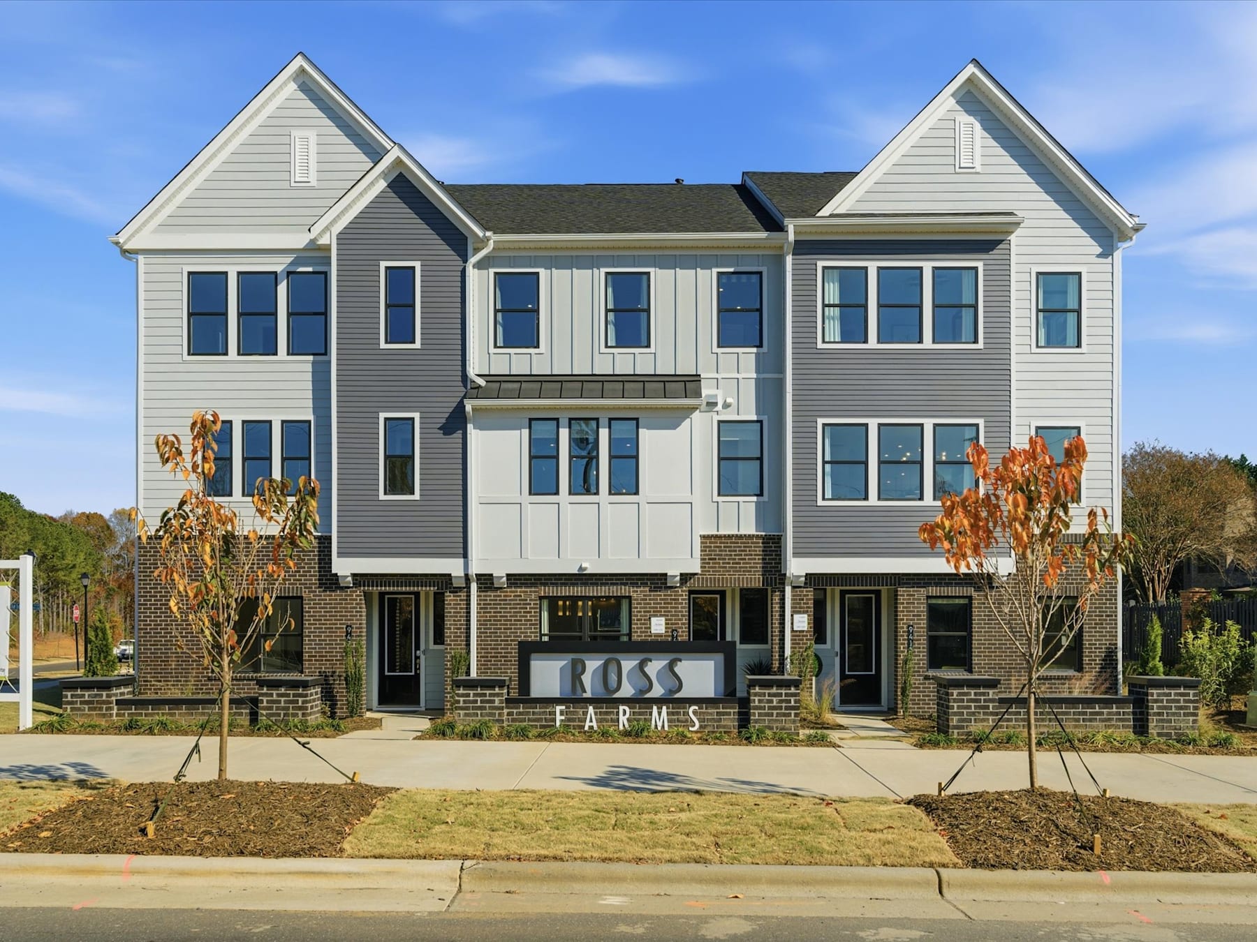 A multi-story townhouse complex with gray siding, black roofs, and large windows, set against a backdrop of blue sky and trees in the foreground.
