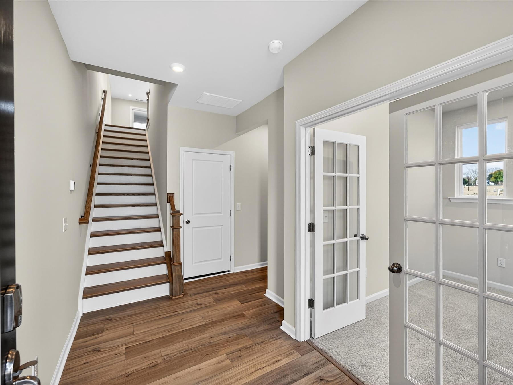 A spacious and well-lit hallway with a wooden staircase leading upstairs, surrounded by white walls and doors, and a hardwood floor.