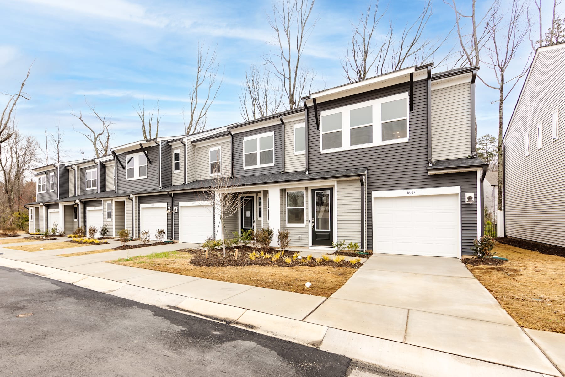A row of modern, two-story townhouses with gray siding and garages, surrounded by bare trees and a paved driveway in the foreground.