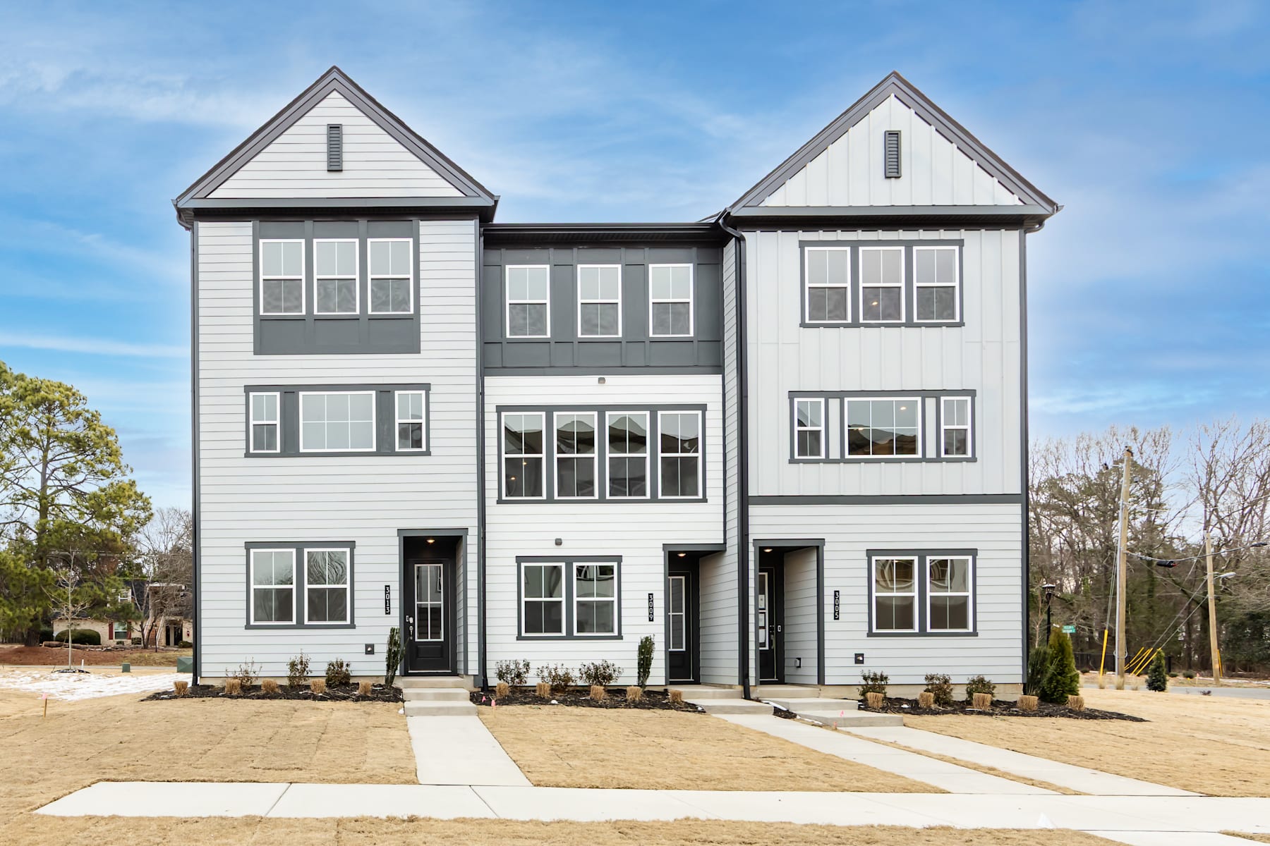 A modern, multi-unit residential building with a gray exterior and white trim, surrounded by a paved walkway and landscaping, set against a backdrop of trees and a clear blue sky.