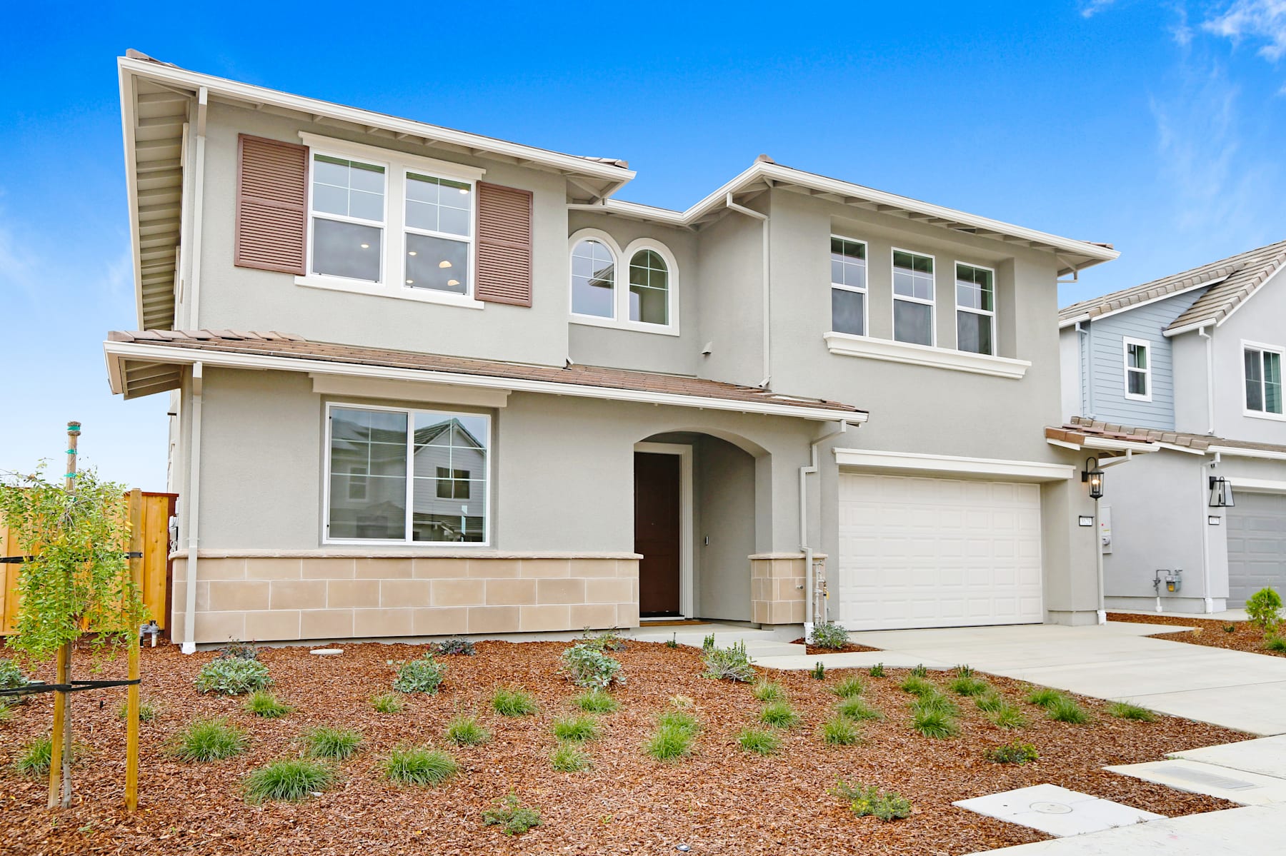 A two-story beige house with a garage, surrounded by a landscaped yard with a grassy area and small plants in the foreground, set against a clear blue sky.