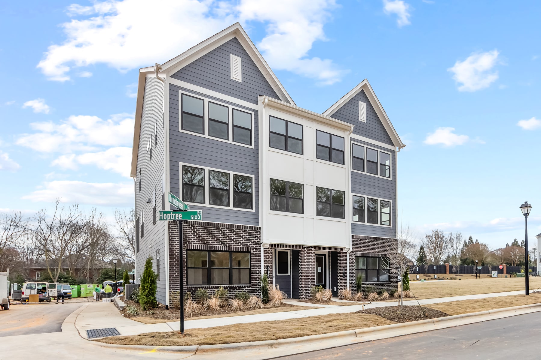 A multi-story residential building with a gray exterior, brick accents, and large windows stands in a landscaped setting with a paved walkway and street lamps in the foreground.