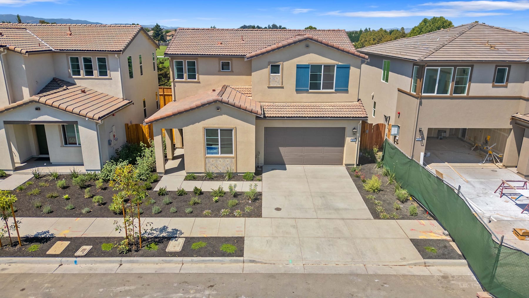 The image shows a residential neighborhood with several two-story houses, each with a garage and a driveway. The houses have a similar architectural style, with tiled roofs and neutral-colored exteriors. The foreground features a paved walkway leading to one of the houses, surrounded by landscaped gardens and trees in the background.