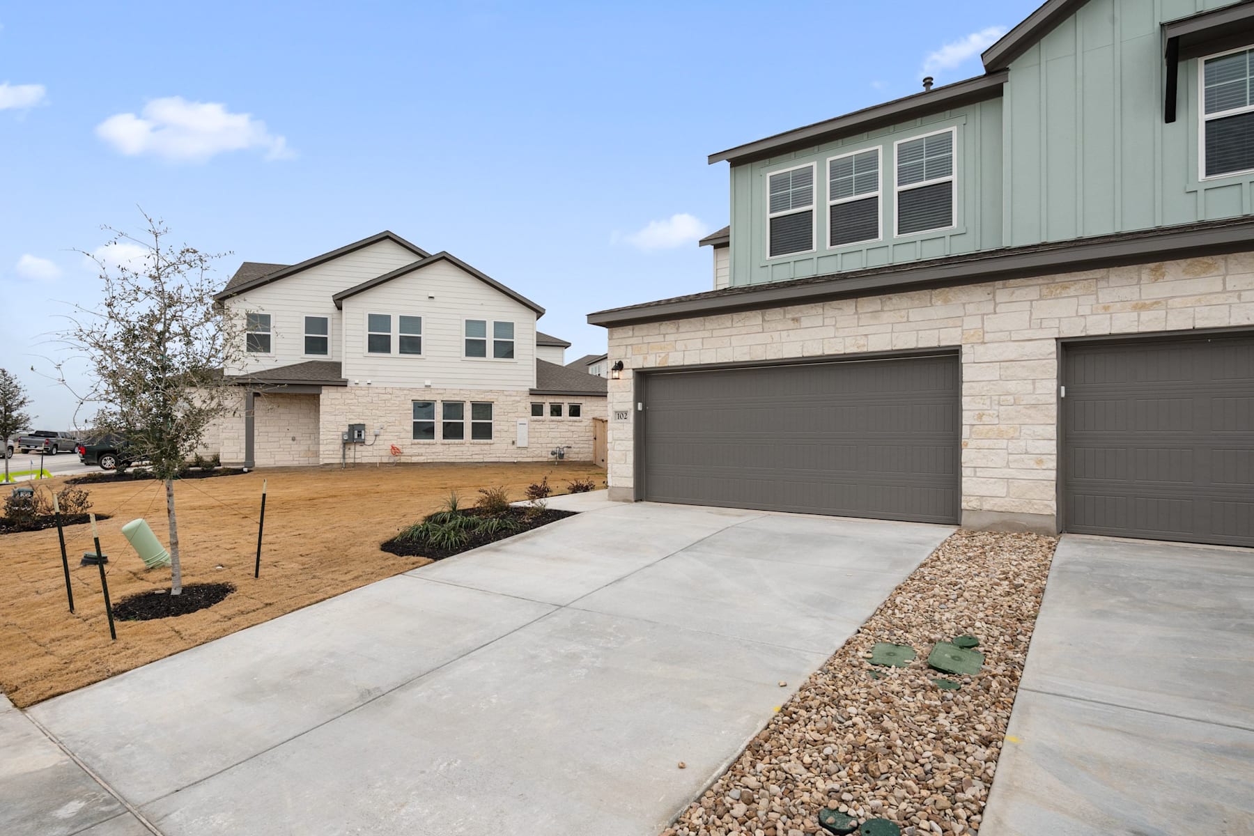 A newly constructed two-story residential building with a garage, surrounded by a paved driveway and landscaped yard with gravel and plants.
