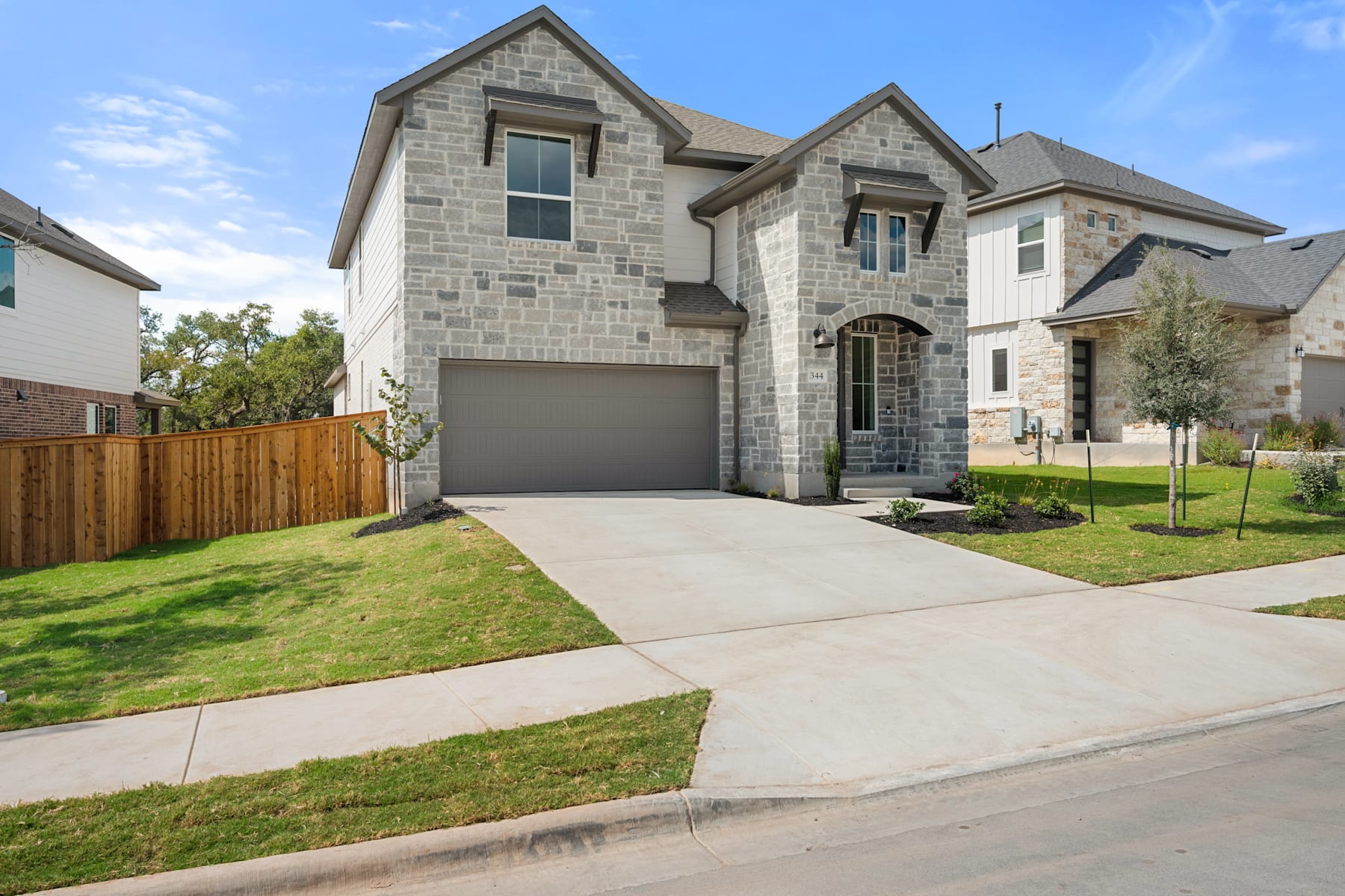 A two-story stone and brick house with a garage, surrounded by a well-manicured lawn and a wooden fence in the foreground, set against a clear blue sky.