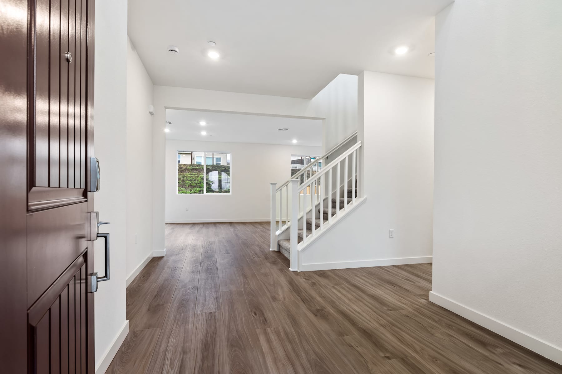 A spacious and well-lit hallway with a wooden floor, white walls, and a wooden staircase leading to the upper level. The hallway appears to be part of a modern and minimalist home interior design.