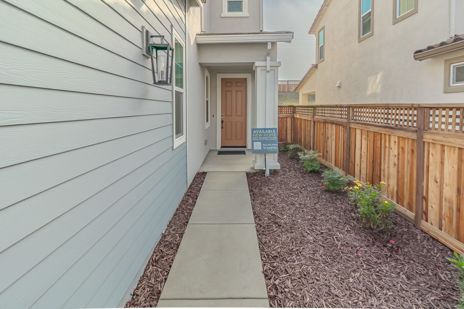 A narrow walkway lined with gravel leads to a wooden door, with a gray siding exterior and a lattice fence on the right side of the image.