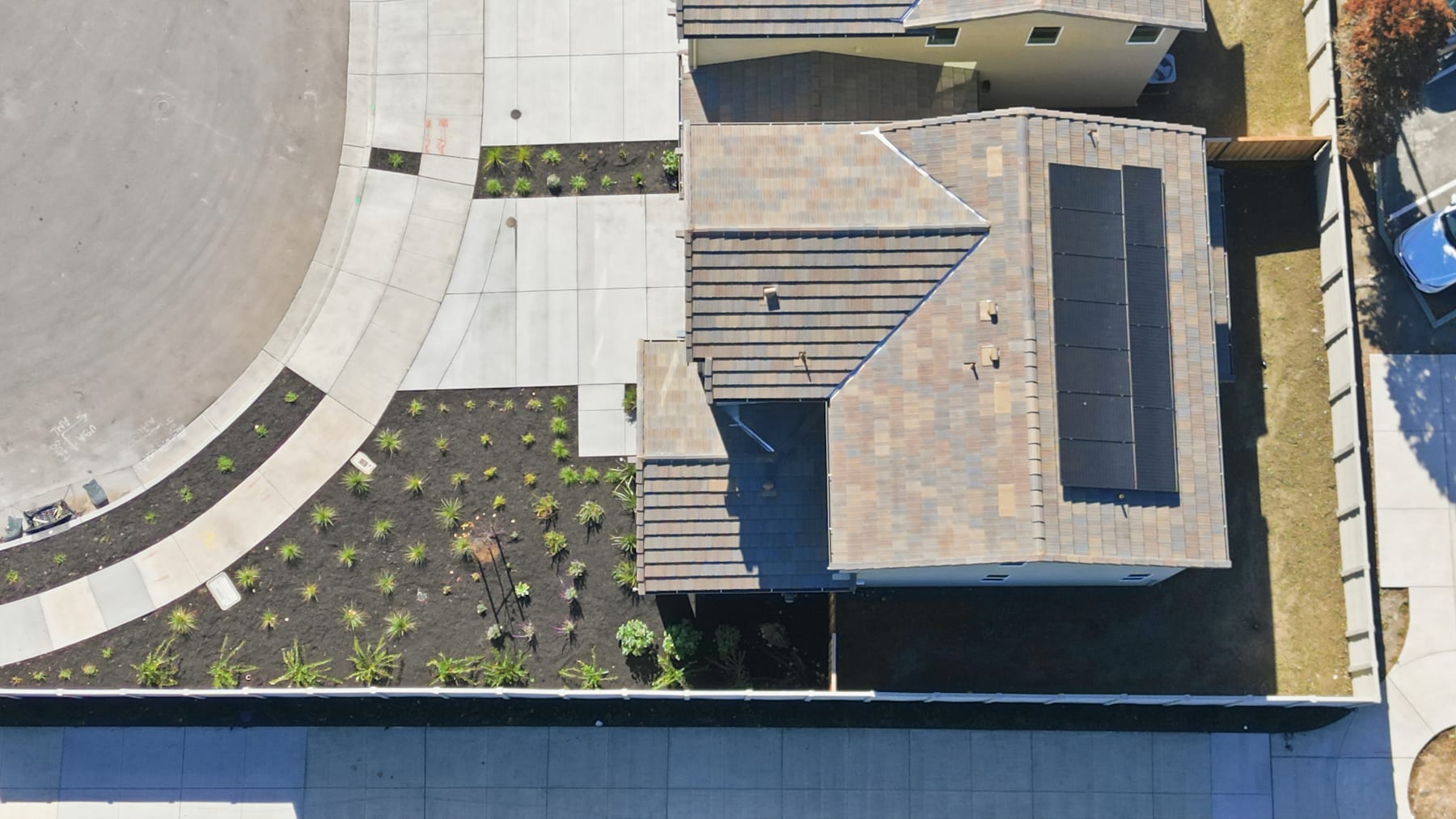 The image depicts a modern, multi-story building with a unique architectural design featuring various geometric shapes, textures, and materials. The foreground includes a landscaped area with small plants or vegetation, while the background showcases other buildings and structures.