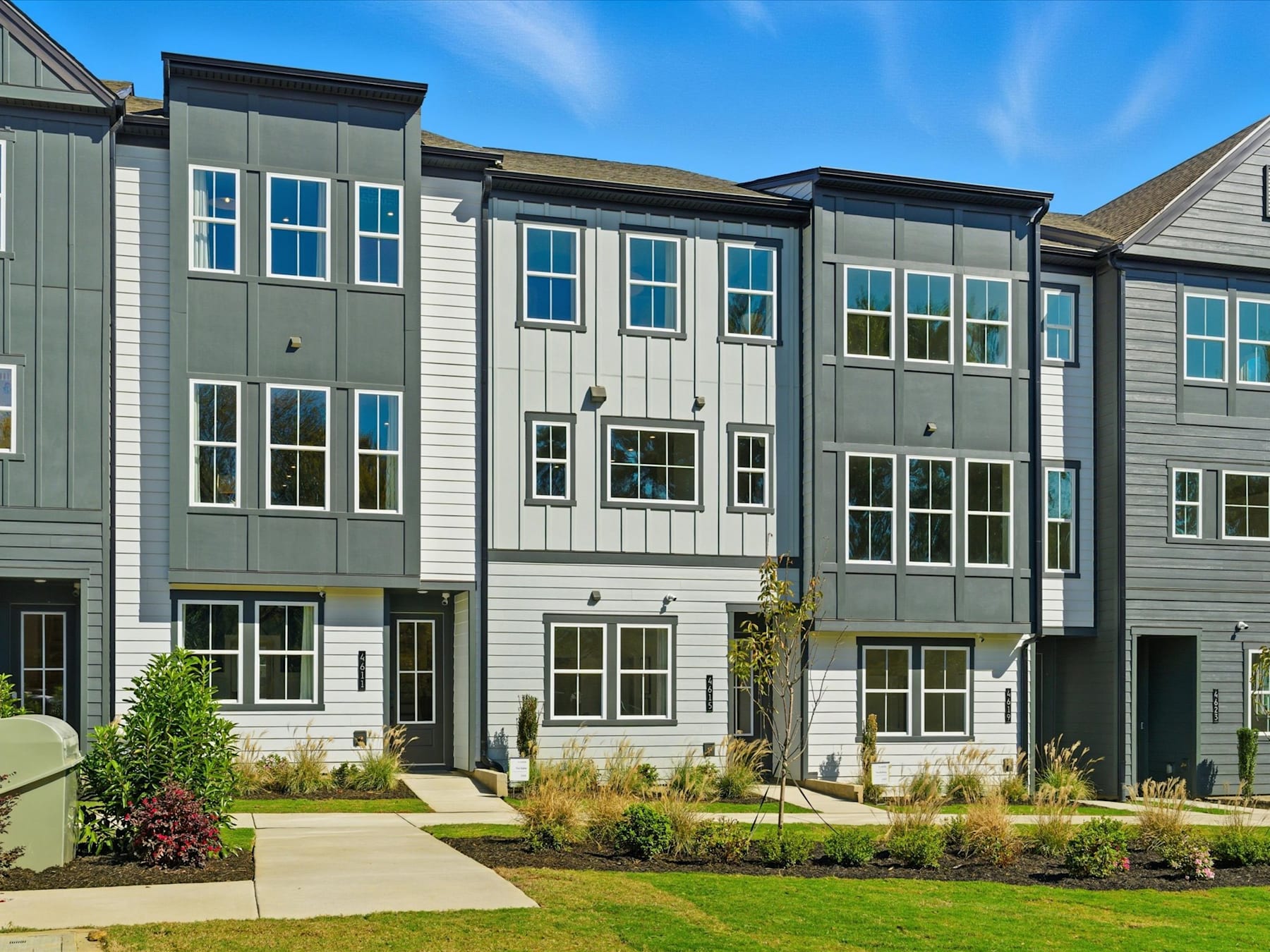The image depicts a row of modern, multi-story townhouses with a well-manicured lawn and landscaping in the foreground, set against a clear blue sky.