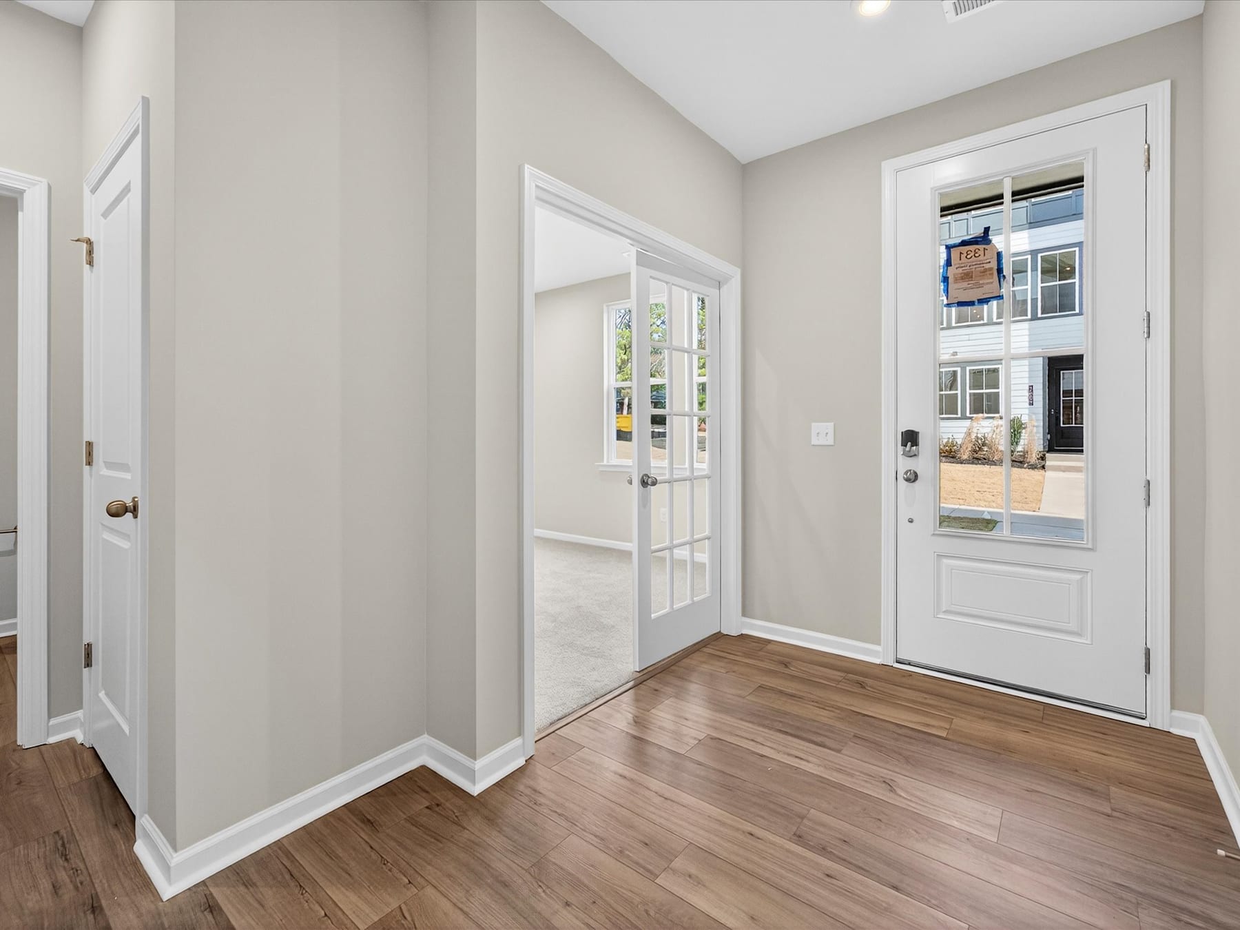 A bright and spacious entryway with white walls, a wooden floor, and a glass-paneled door leading to the exterior.