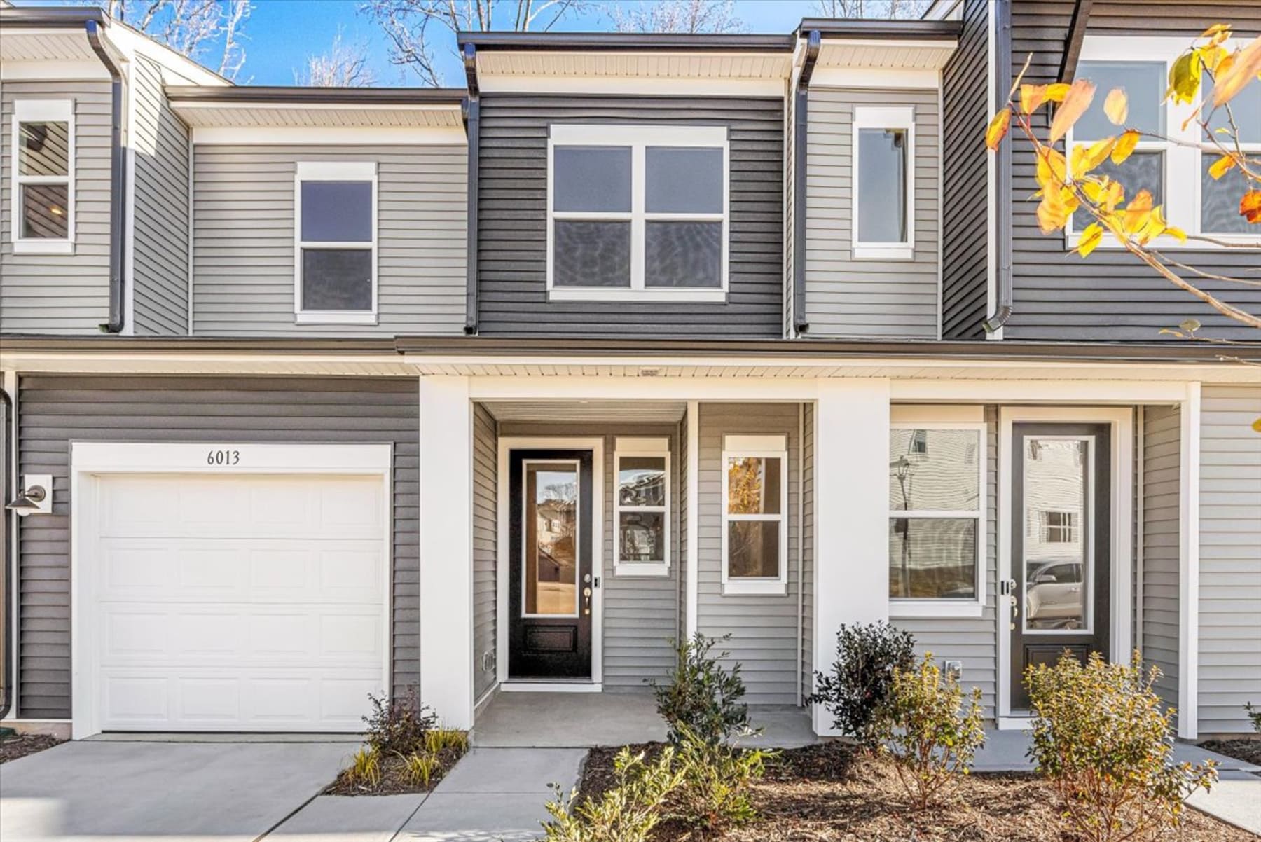 A two-story townhouse with a white garage door, gray siding, and a covered porch surrounded by landscaping against a backdrop of a clear blue sky and bare trees.