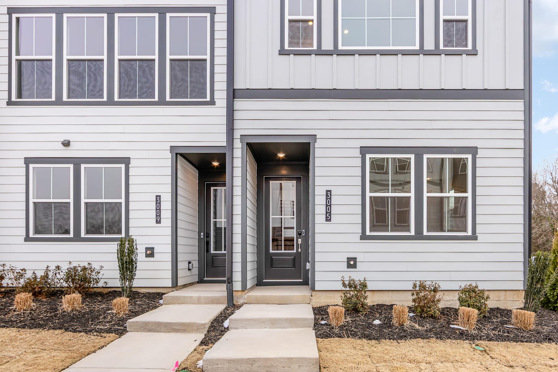 A modern, two-story residential building with a gray exterior, large windows, and a paved walkway leading to the front entrance.