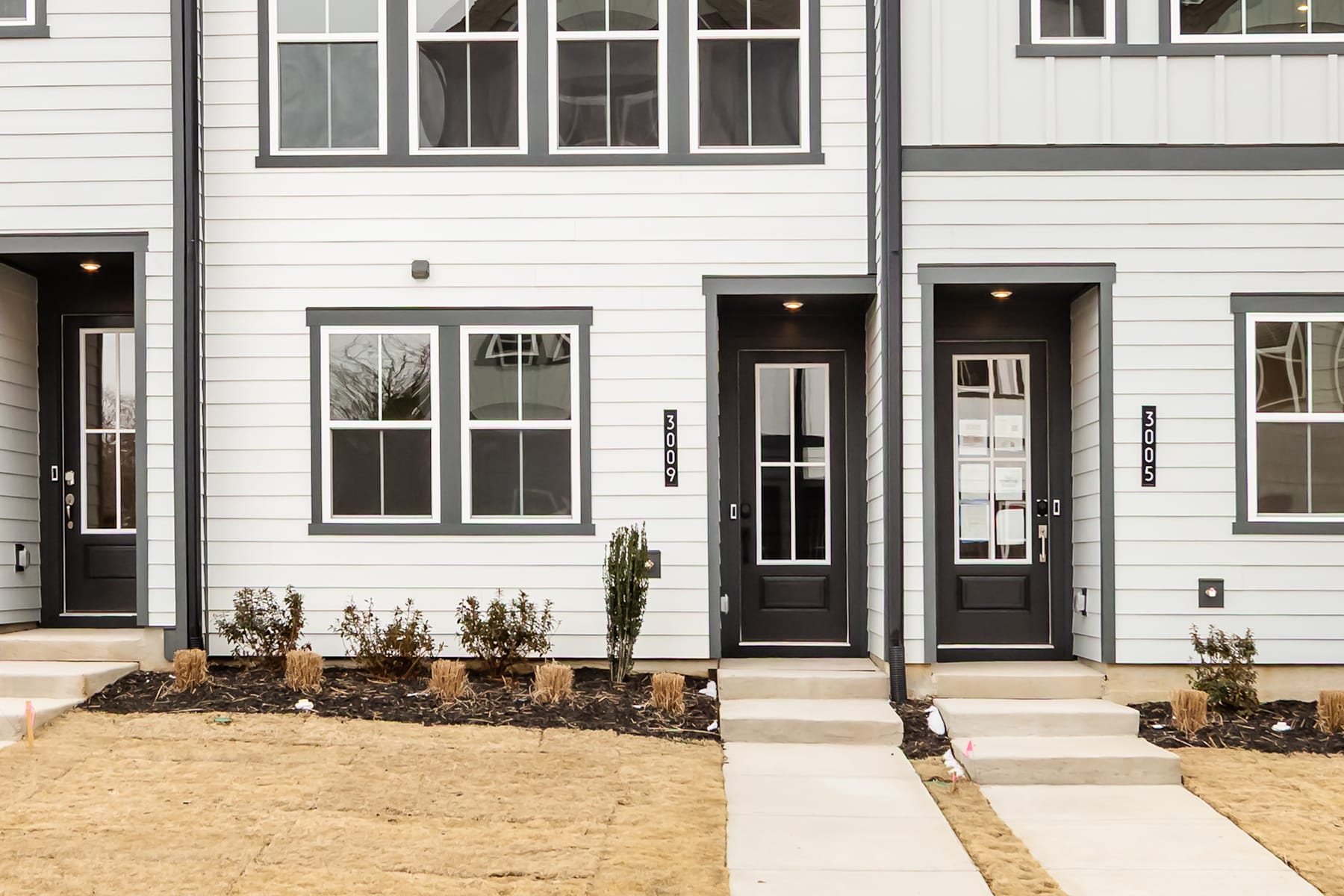 The image shows a modern townhouse with a gray exterior, featuring multiple entrances and windows. The foreground includes a paved walkway leading to the front doors, surrounded by small plants and landscaping.