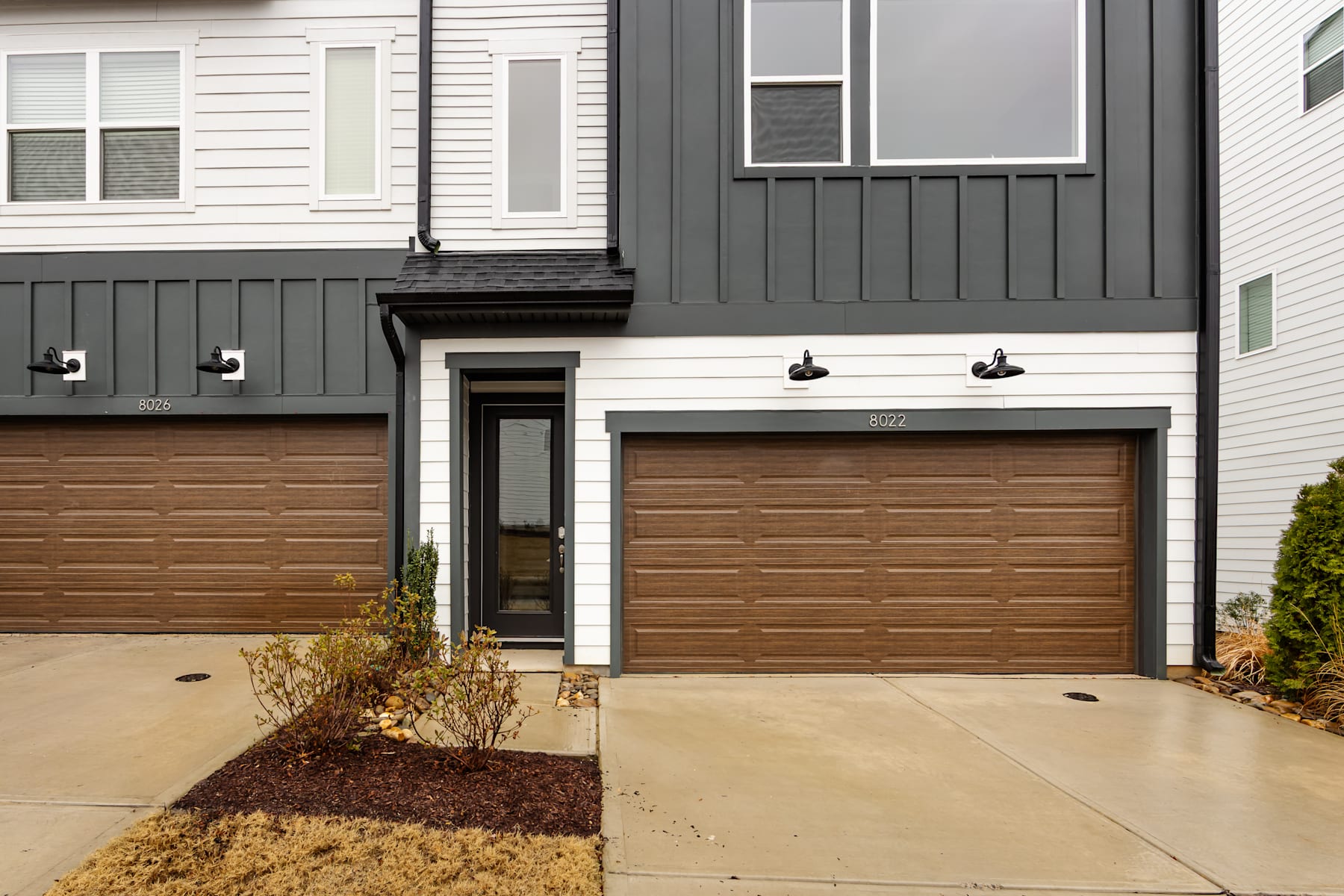 A modern townhouse with a wooden garage door, surrounded by a concrete walkway and landscaping in the foreground.