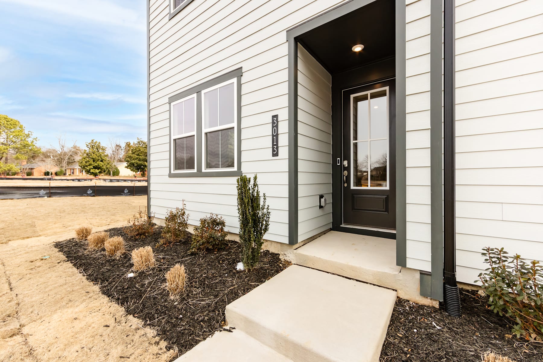 A modern, gray-sided house with a paved walkway leading to the front door, surrounded by landscaped beds with small plants and shrubs.
