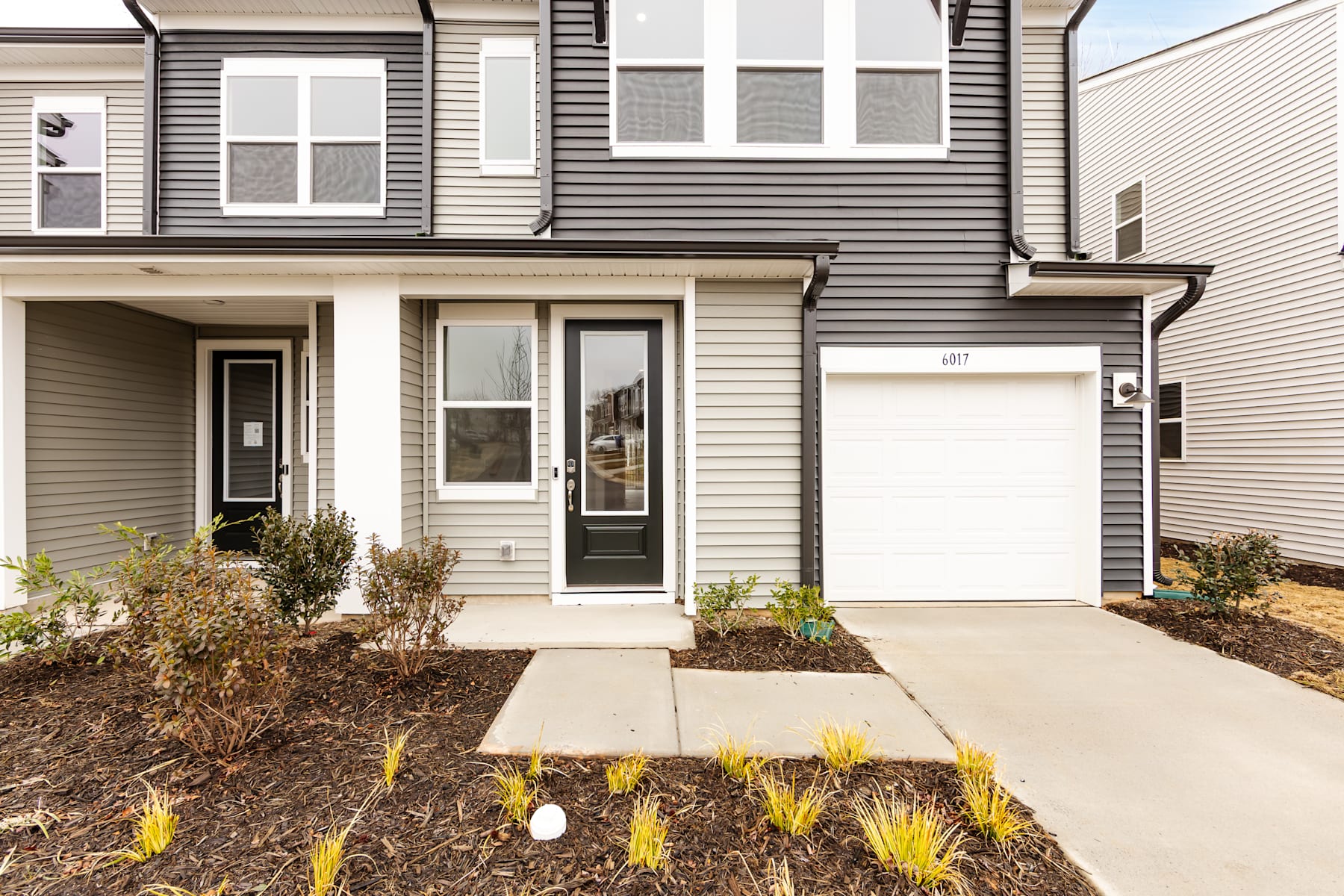 A two-story townhouse with gray siding, a garage door, and a paved walkway leading to the front entrance, surrounded by landscaped vegetation.