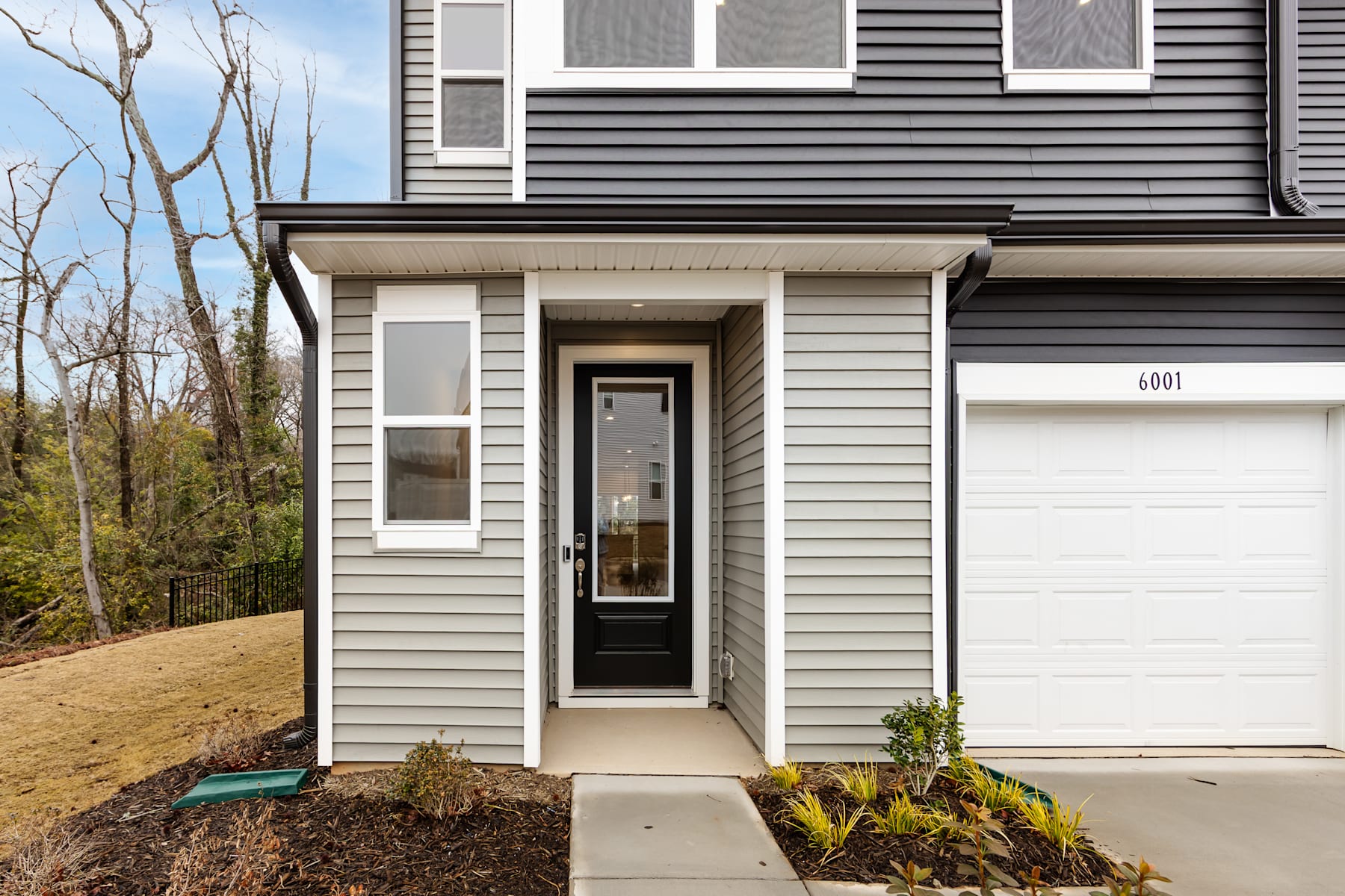 A modern two-story house with gray siding, a garage door, and a small landscaped area in the foreground, set against a backdrop of trees and a clear sky.