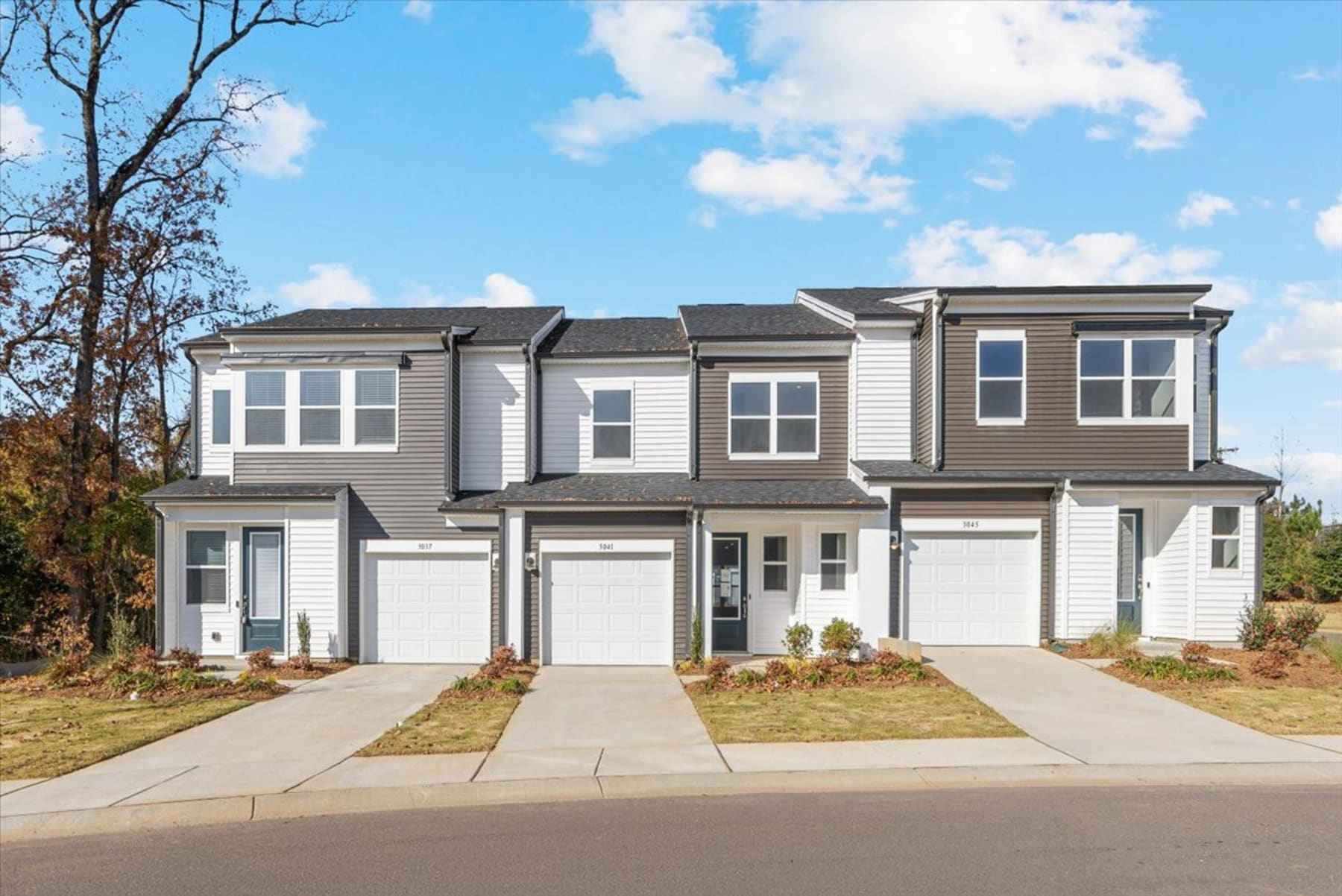 A modern, multi-unit residential building with a gray exterior, white trim, and a well-landscaped front yard, set against a backdrop of blue sky with scattered clouds.
